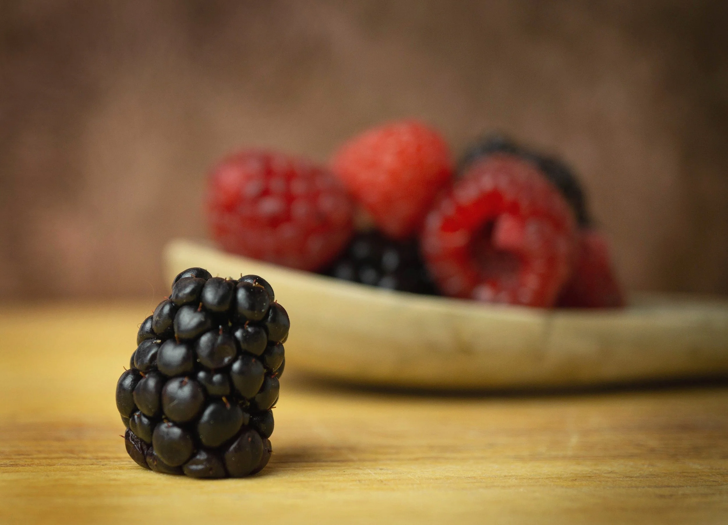Close-up of a blackberry on a wooden surface with raspberries in a blurred background.