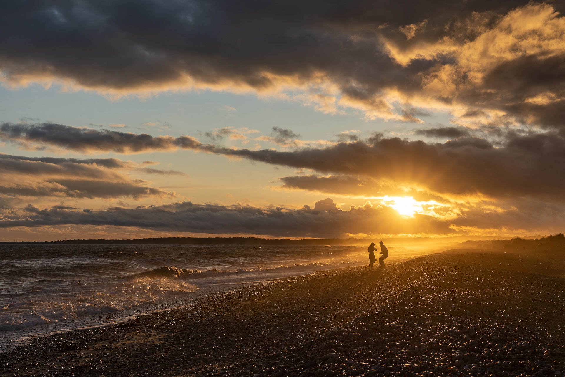 Two people on a rocky beach at sunset with dramatic clouds in the sky.
