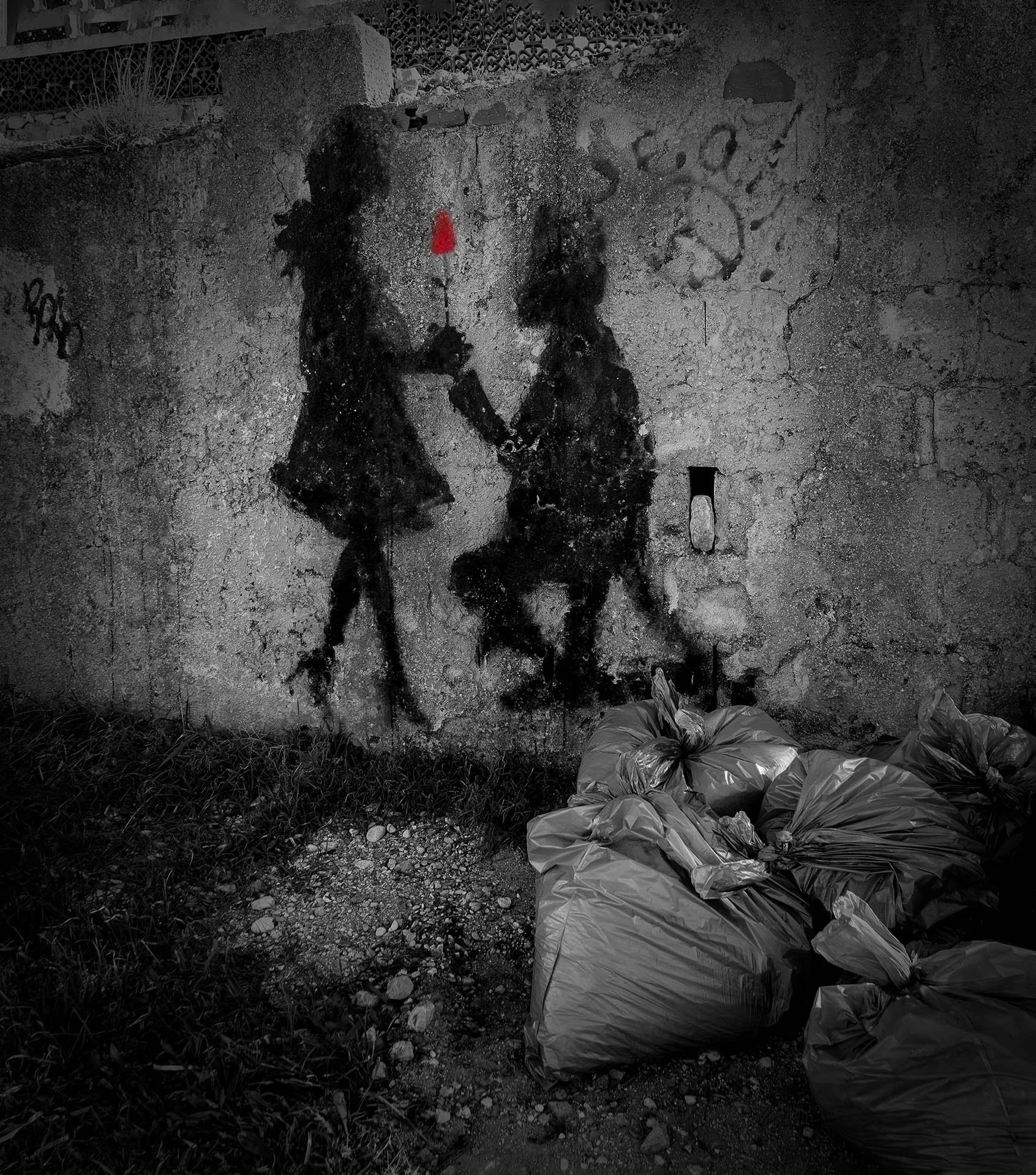 A black and white graffiti on a wall shows a silhouette of a person on one knee offering a red rose to another standing person. In front of the wall, there are several garbage bags on the ground.