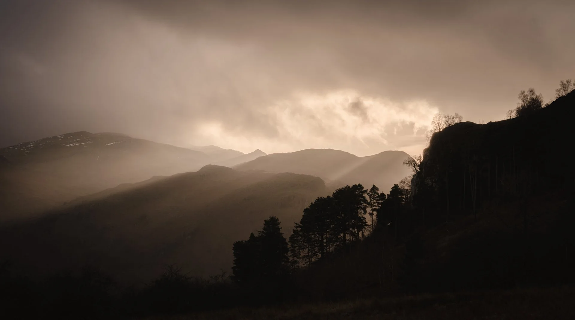 Silhouetted mountain landscape at sunset with sunbeams filtering through clouds, casting a warm glow over ridges and trees.