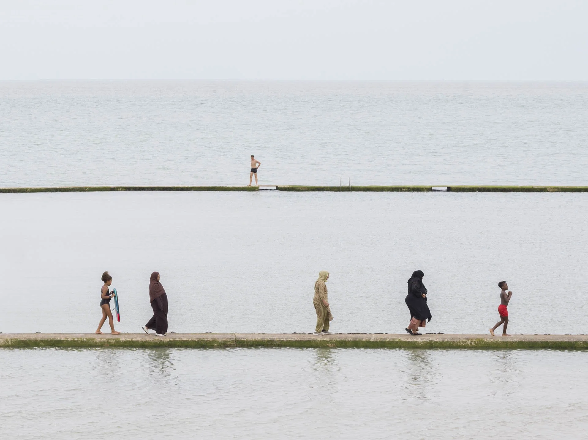 People walking on a narrow path by the sea with a person standing in the background.
