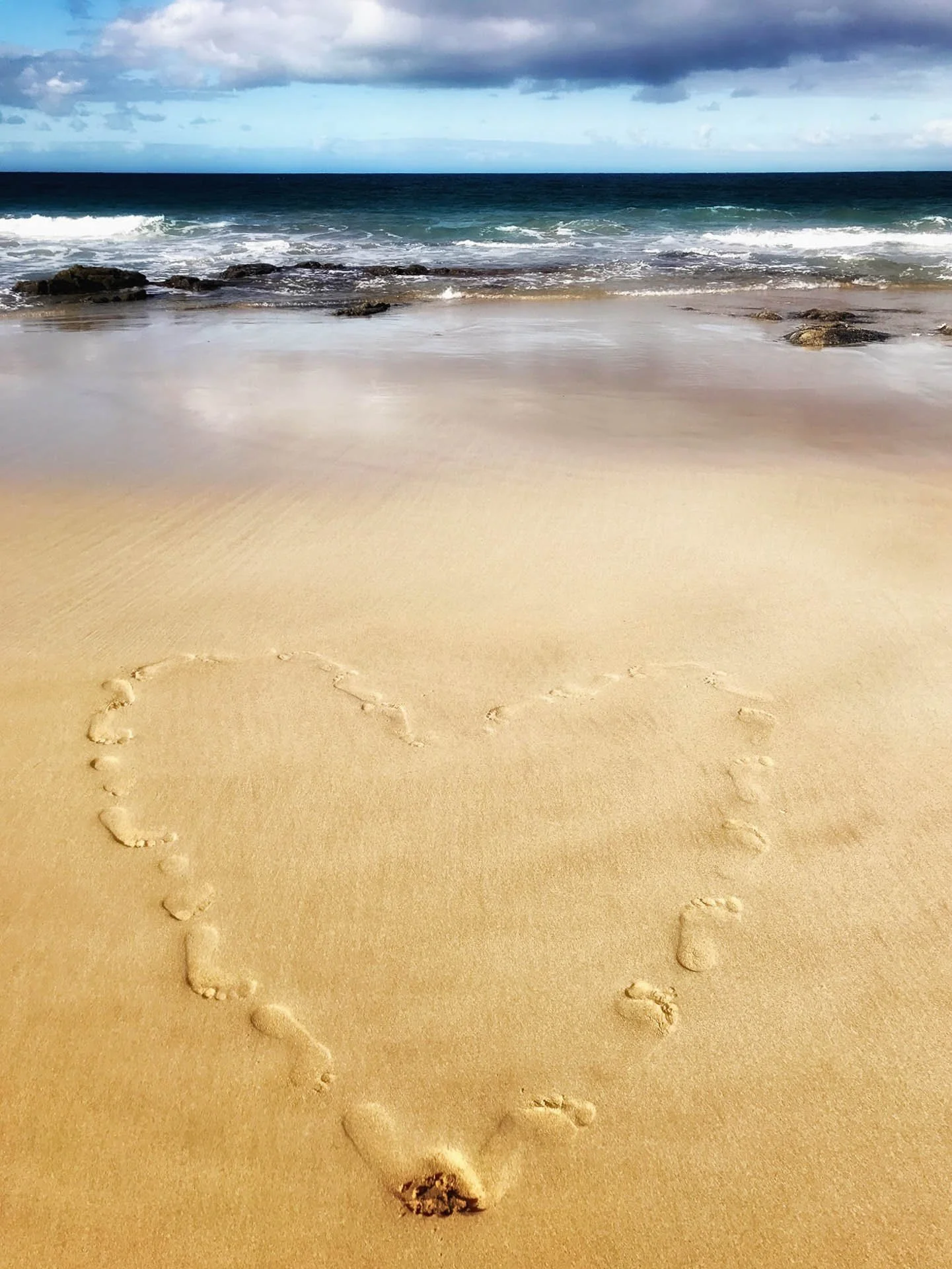 Heart shape drawn with footprints on a sandy beach by the ocean.