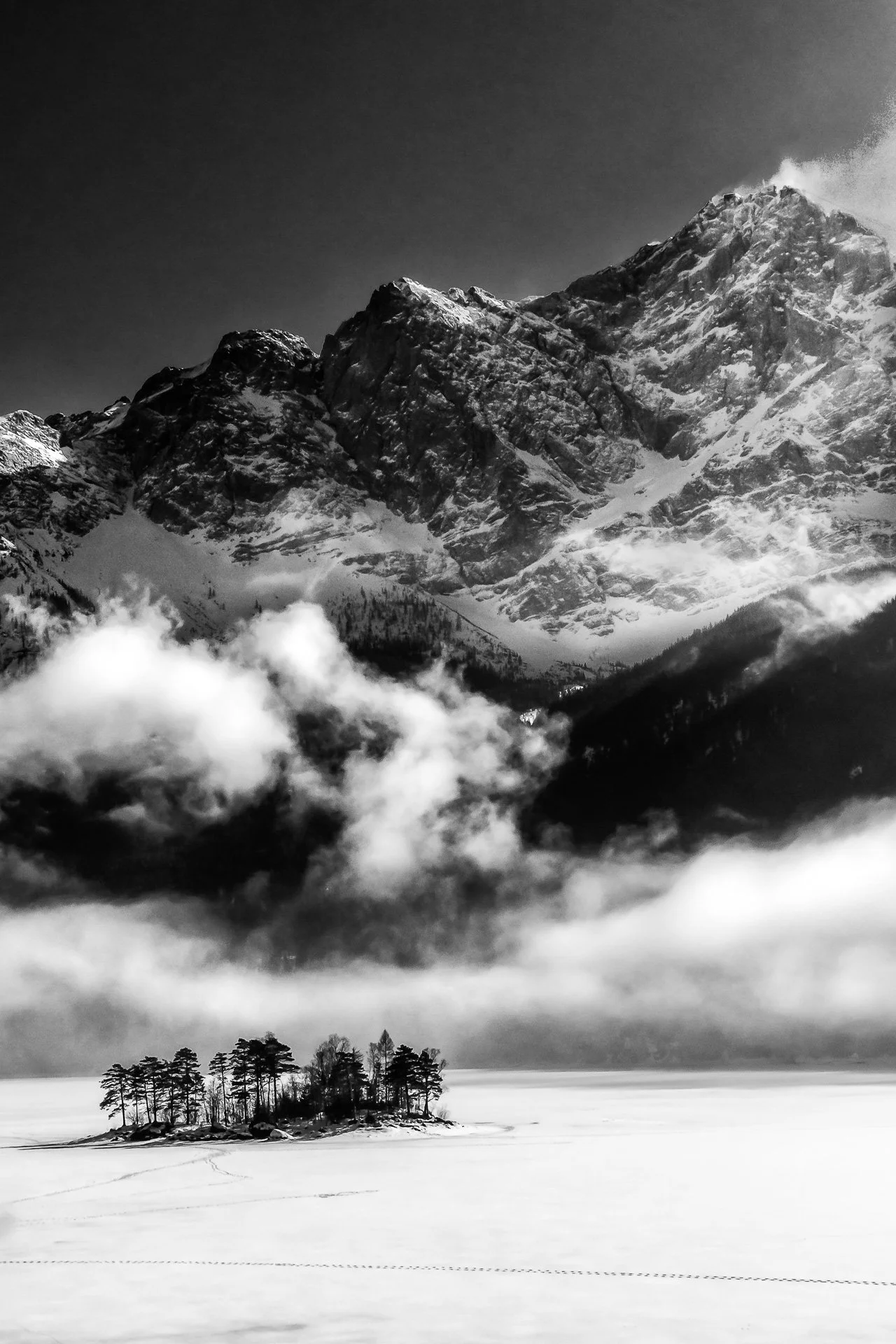 Black and white photo of a snowy mountain landscape with clouds and a small island of trees on a frozen lake.