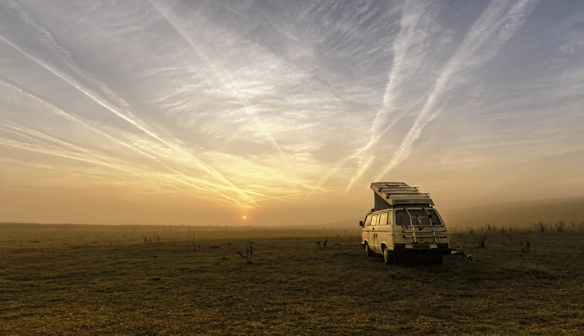 Camper van parked on a vast field at sunset with a dramatic sky and visible contrails.
