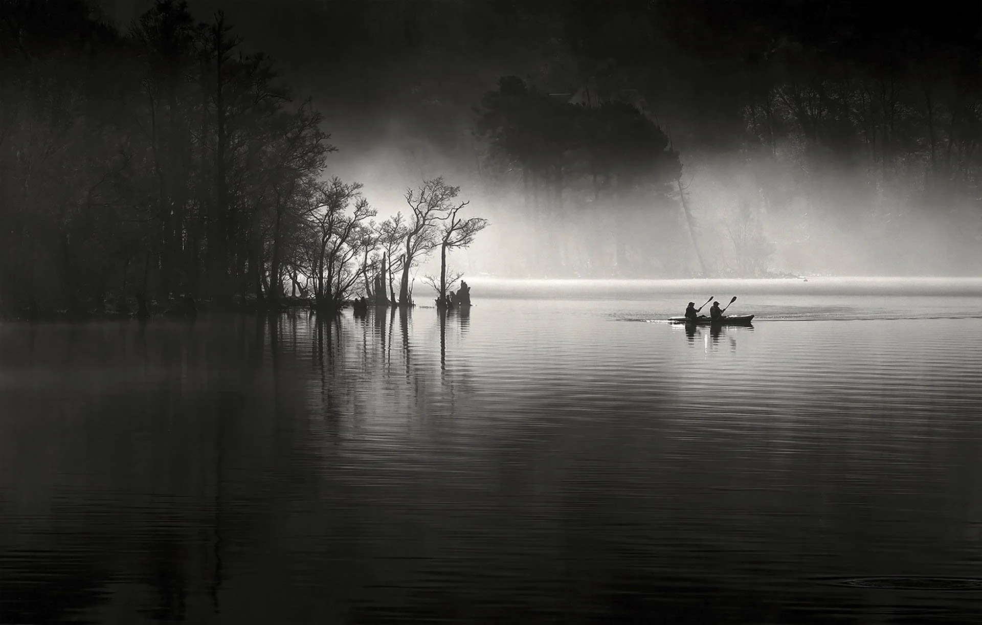 Two people canoeing on a misty lake surrounded by trees, captured in black and white.
