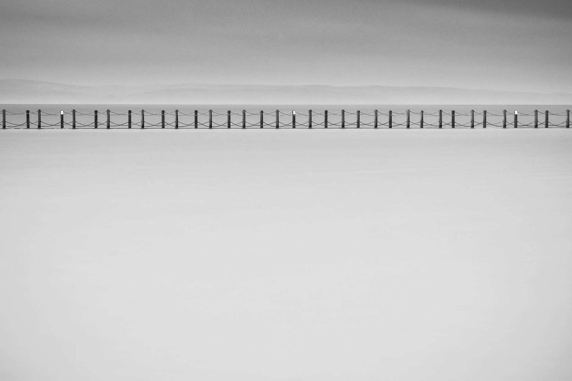 Minimalist black-and-white photo of a long pier with a railing over calm water.