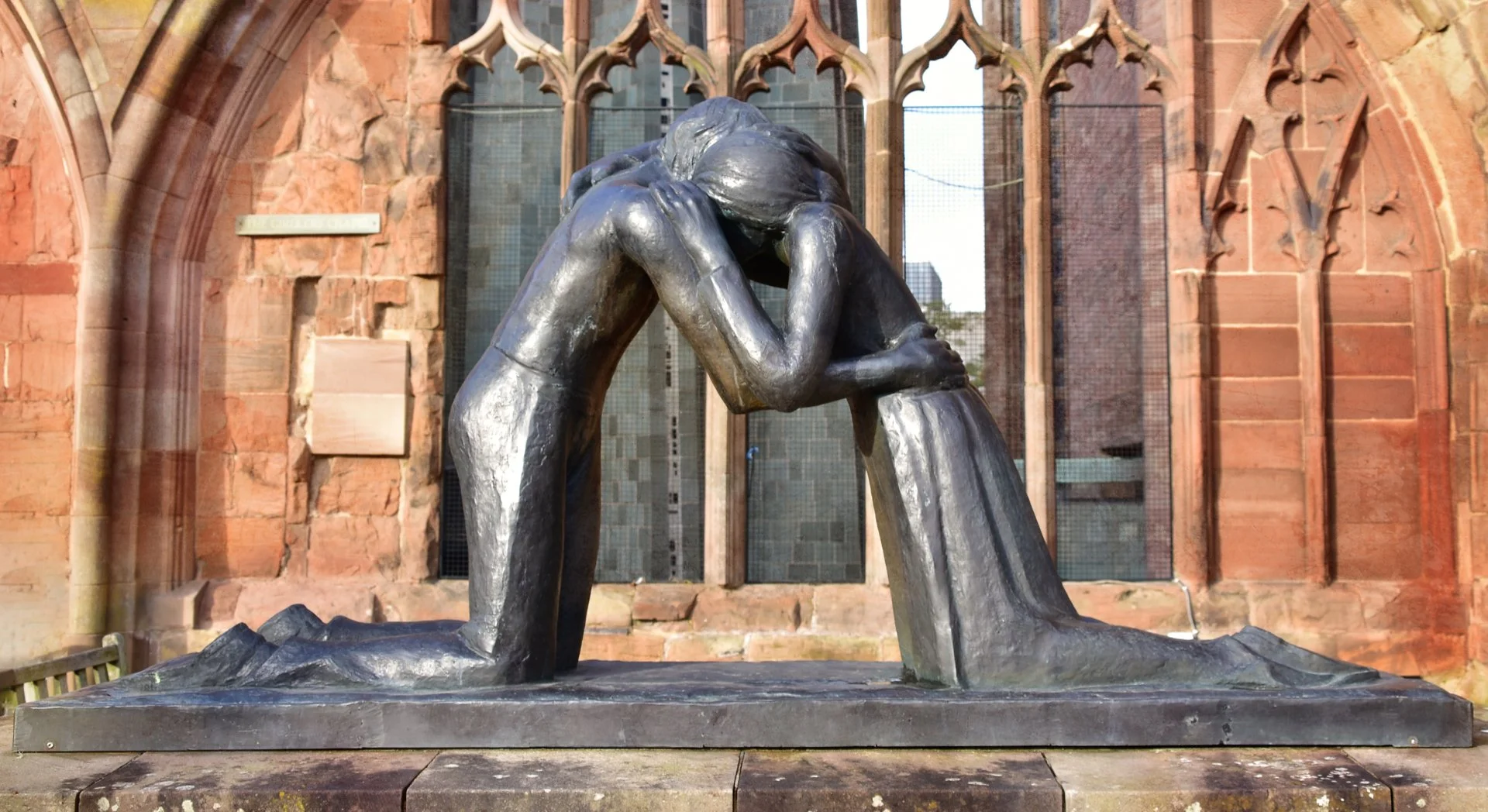 Bronze sculpture of two kneeling figures embracing, set against a stone wall backdrop with arched openings.