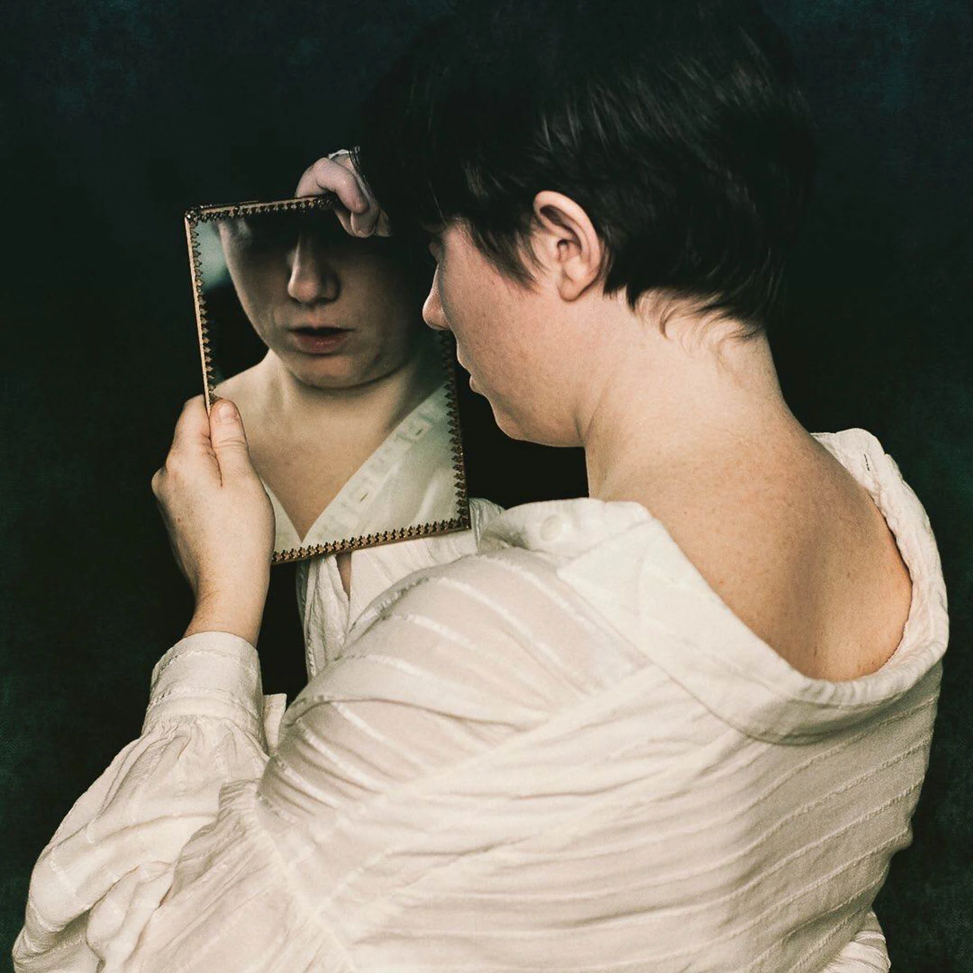Person holding a mirror reflecting their face, wearing a white shirt, against a dark background.