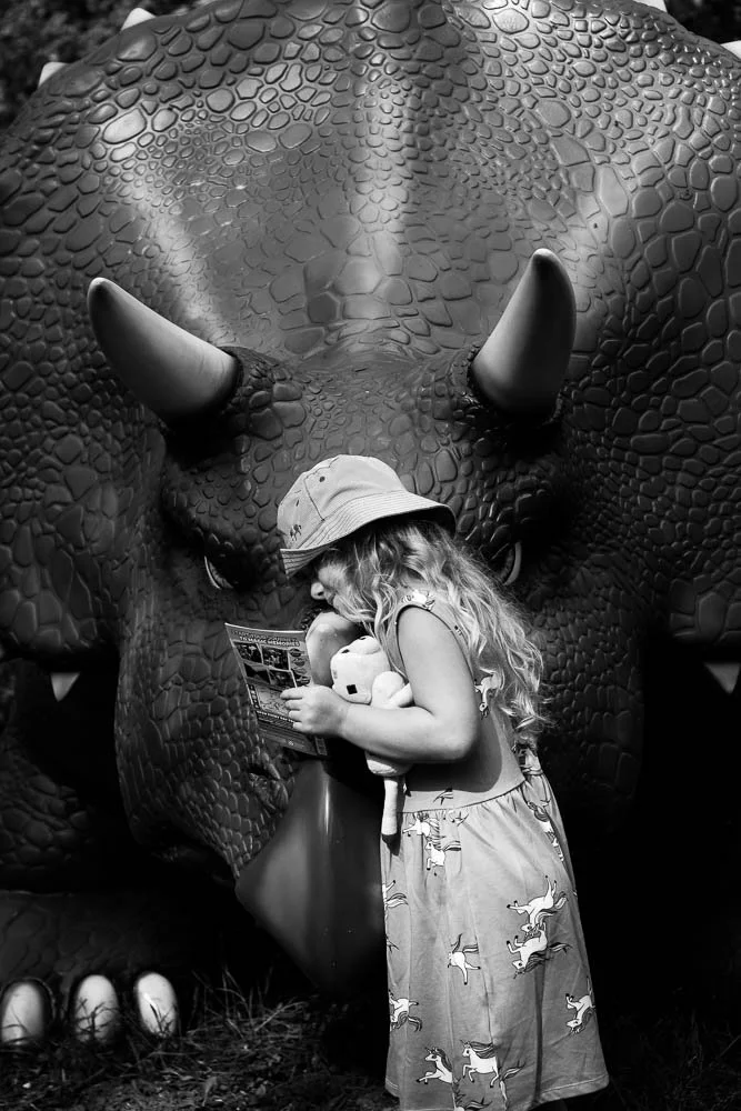 A young child with long hair, wearing a sun hat and a dress with a horse pattern, stands in front of a large triceratops sculpture. The child is holding a toy animal and appears to be reading an informational pamphlet. The scene is captured in black