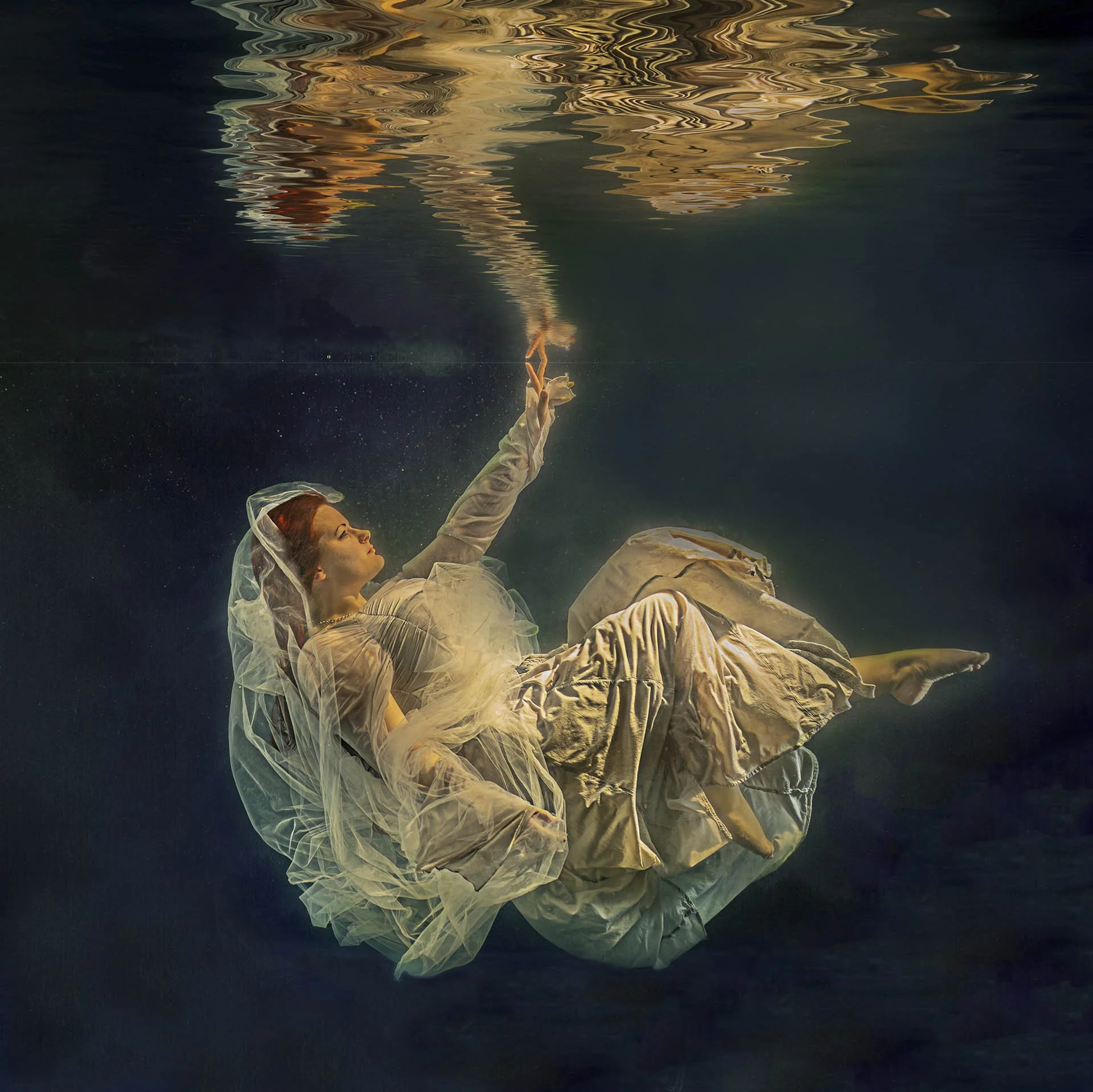 Underwater photo of a woman in a white dress reaching towards the surface, surrounded by water reflections.