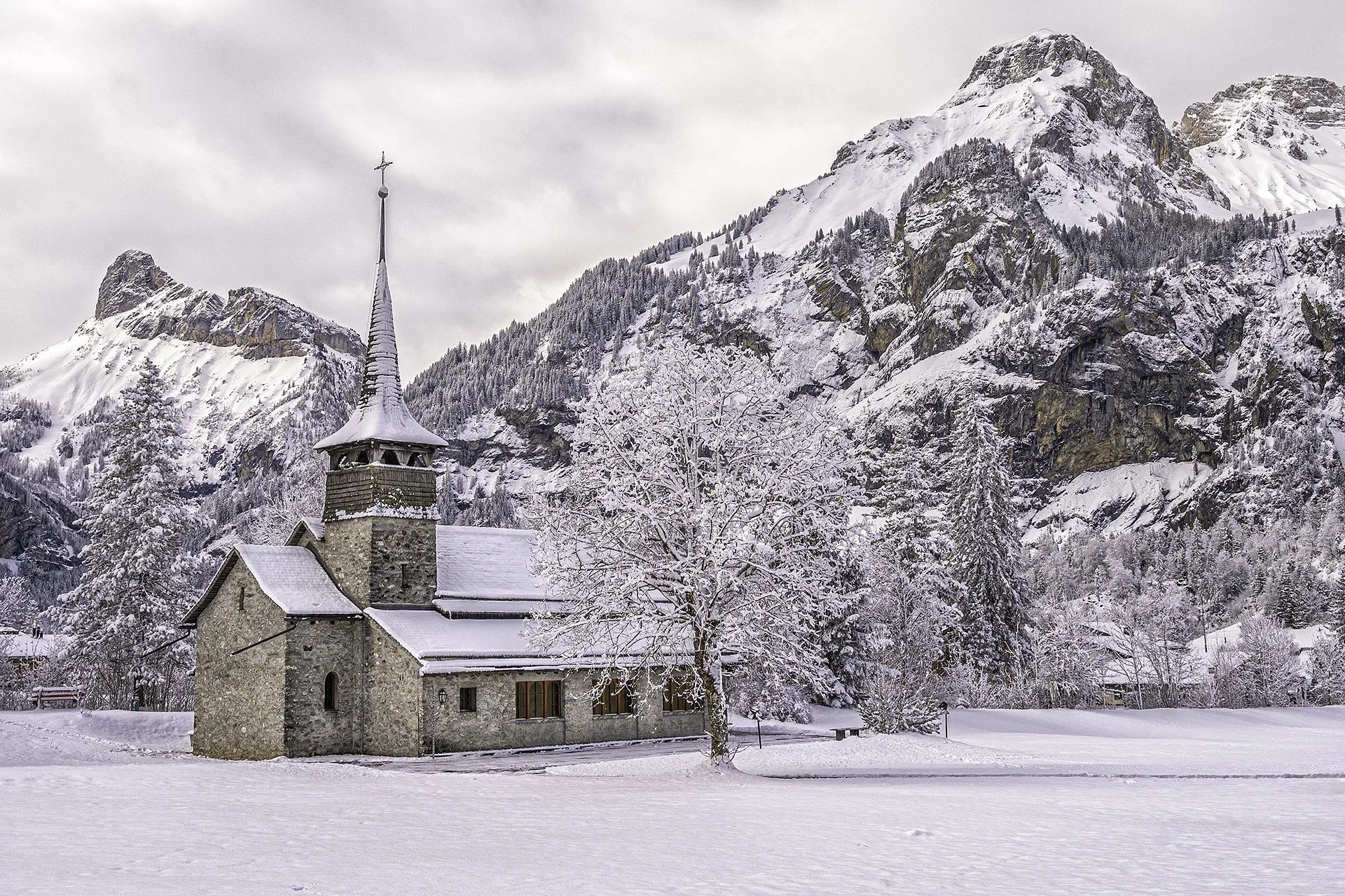 Snow-covered stone church with a tall steeple set against a backdrop of snow-capped mountains and trees.