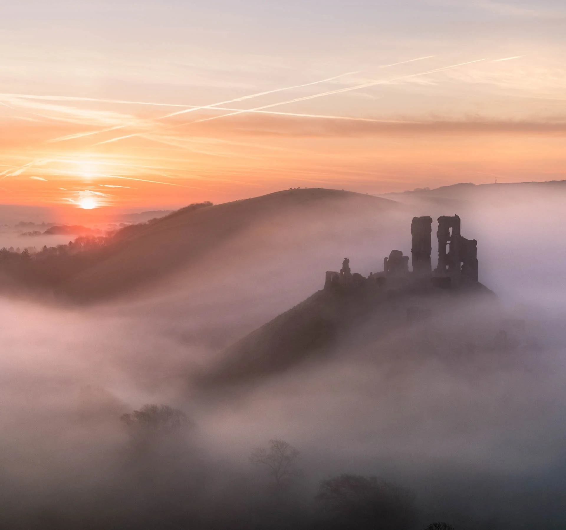 Sunrise over a misty landscape with a silhouette of an old castle on a hill, surrounded by foggy hills and scattered trees.