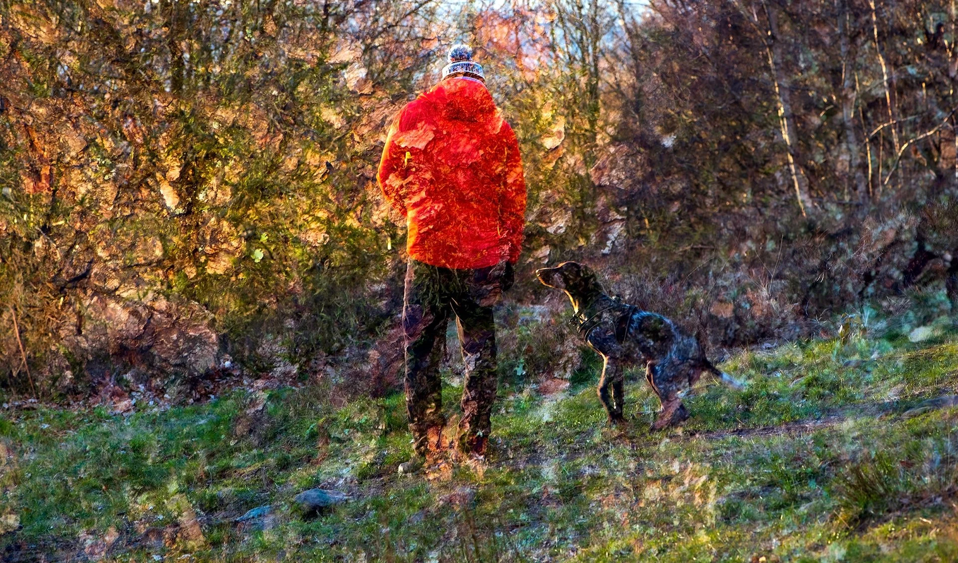 Double exposure image of a person in a bright red jacket standing outside with a dog, surrounded by autumn trees and grass.