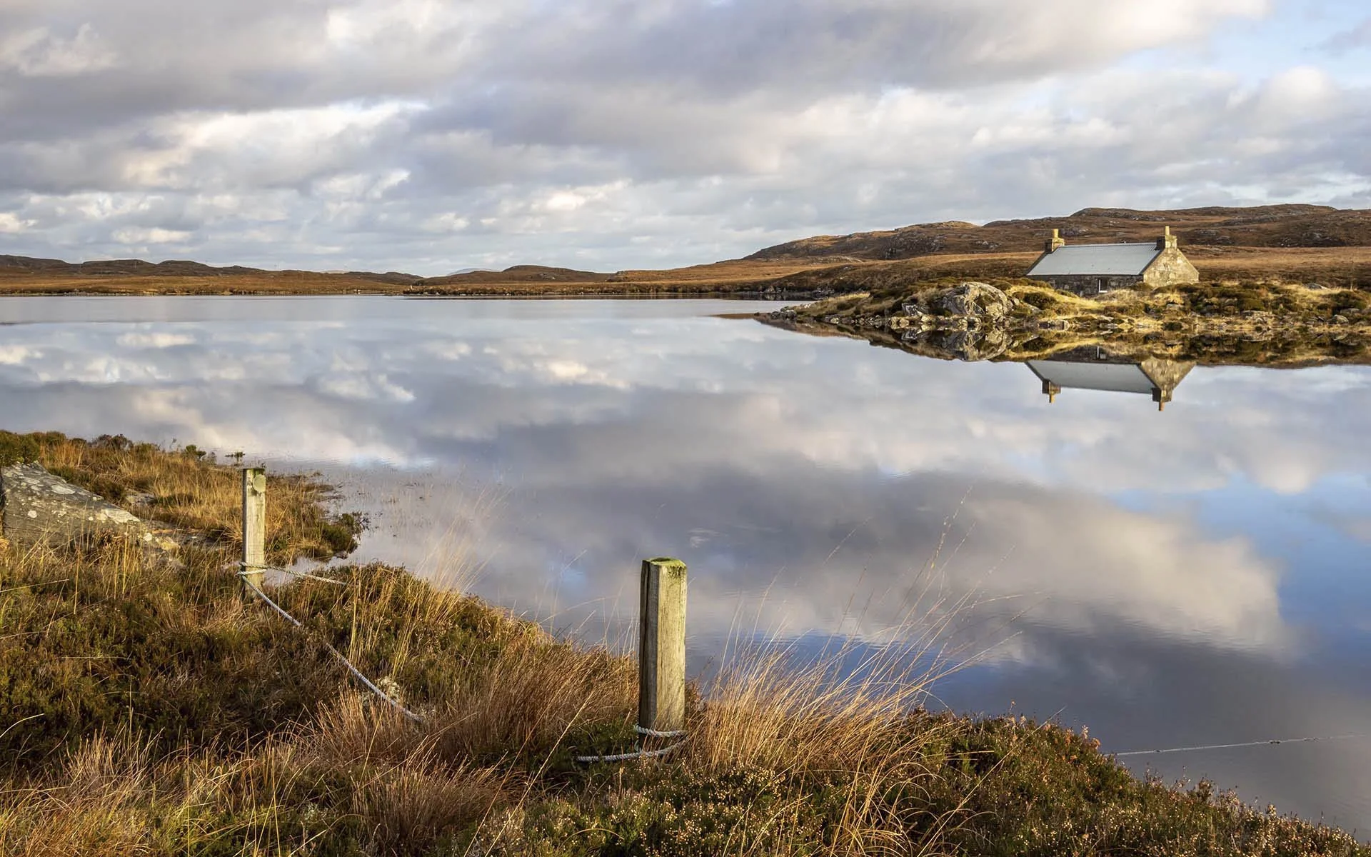 Scenic view of a serene lake with reflections of clouds and a small rustic house on the shoreline, surrounded by grassy terrain and hills.