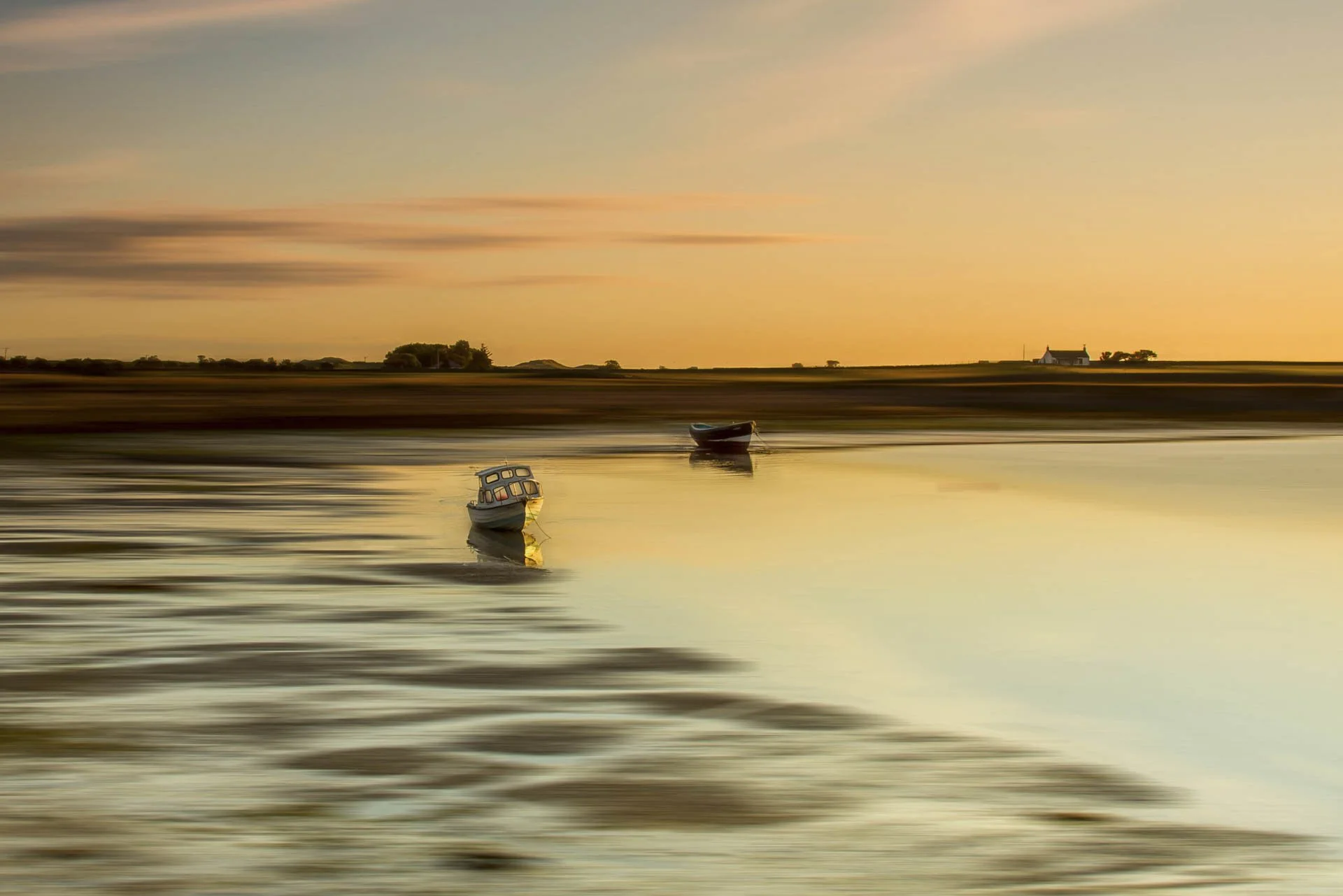Two boats on tranquil water at sunset with a rural landscape and small house in the background.