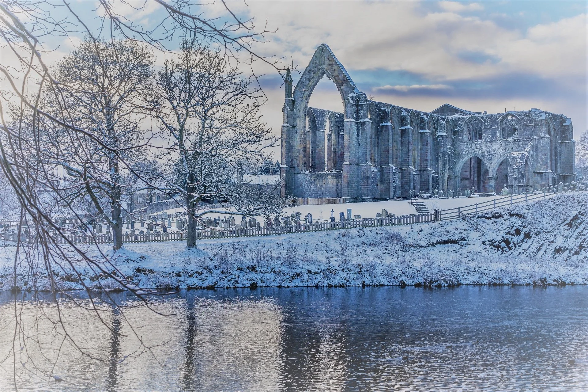 Snow-covered ruins beside a river, with bare trees and a cloudy sky in the background.