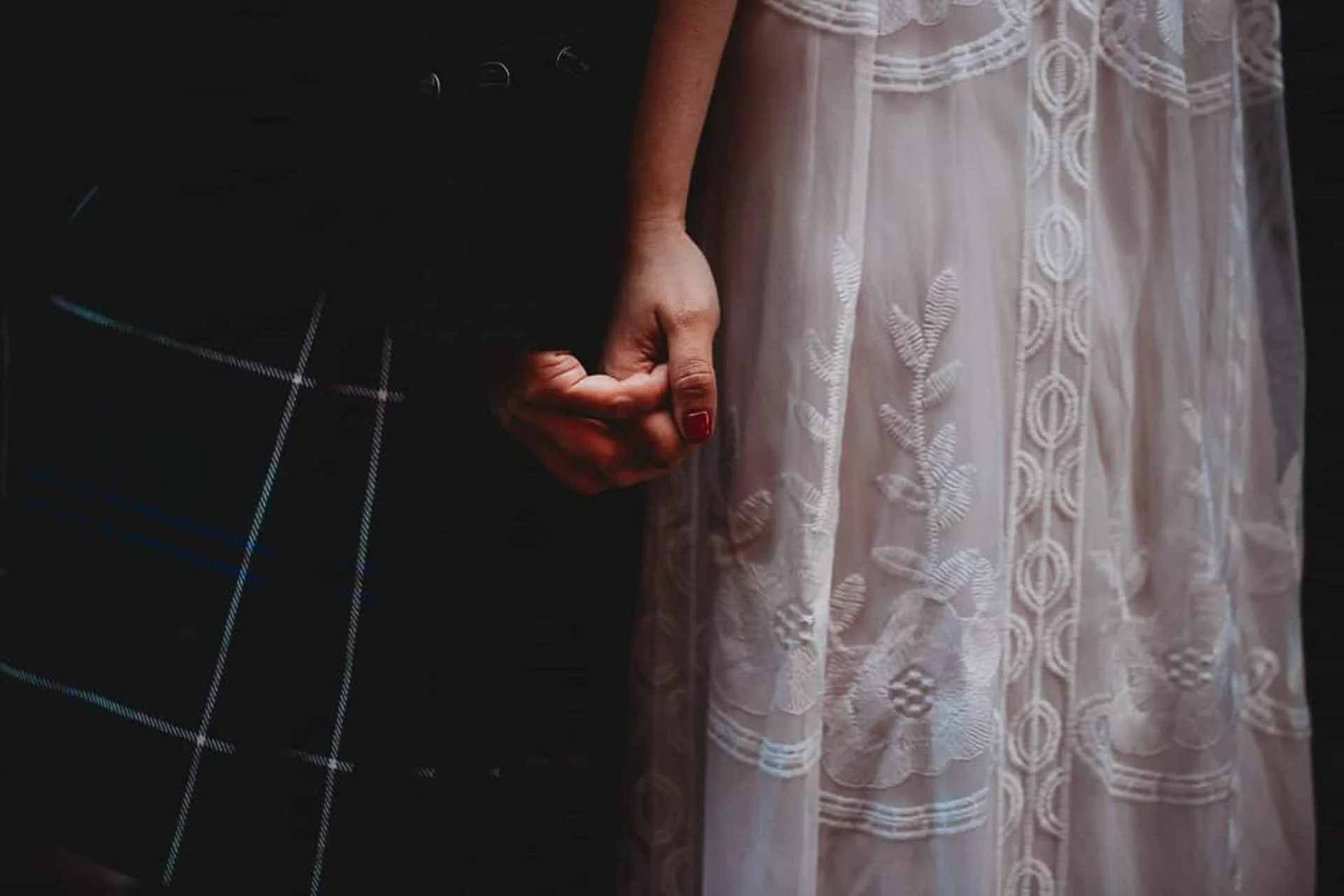 A close-up of a couple holding hands. One person is wearing a dark plaid garment, while the other is wearing a white, lace-detailed dress.