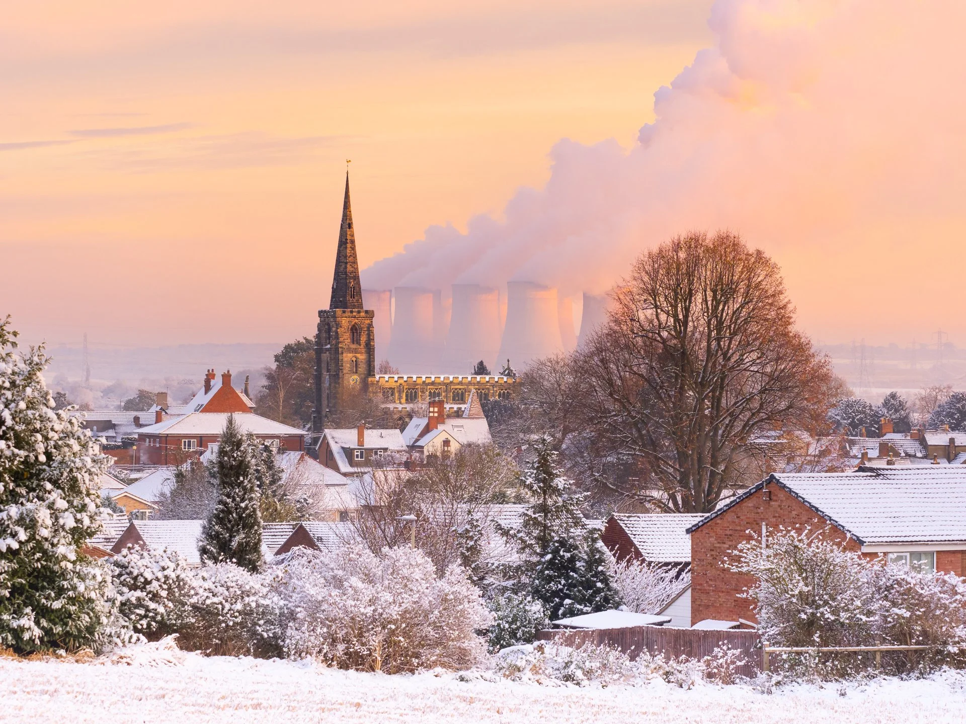 Snow-covered village with church spire and cooling towers in the background at sunrise.