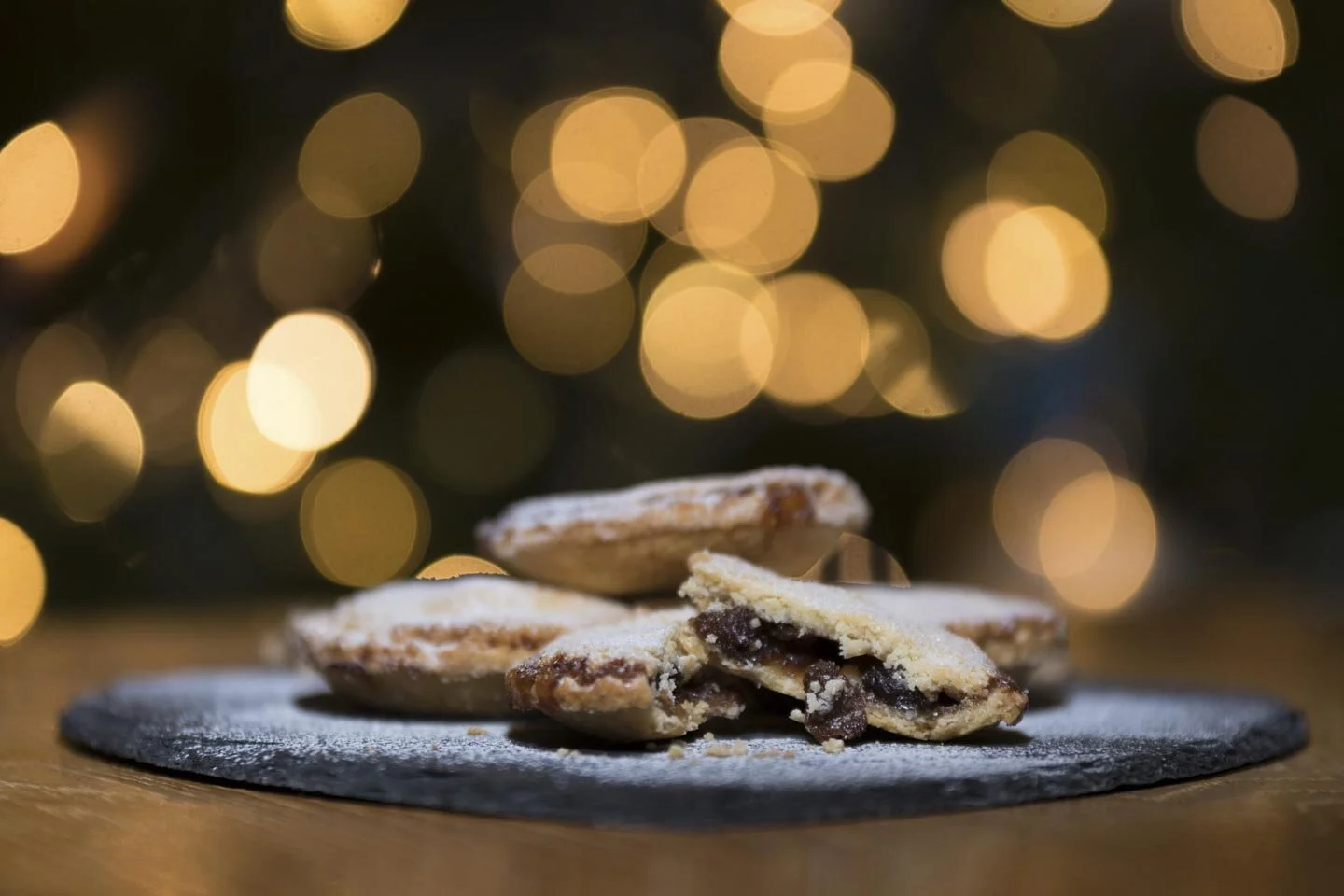Close-up of mince pies with powdered sugar on a plate, blurred festive lights in the background.