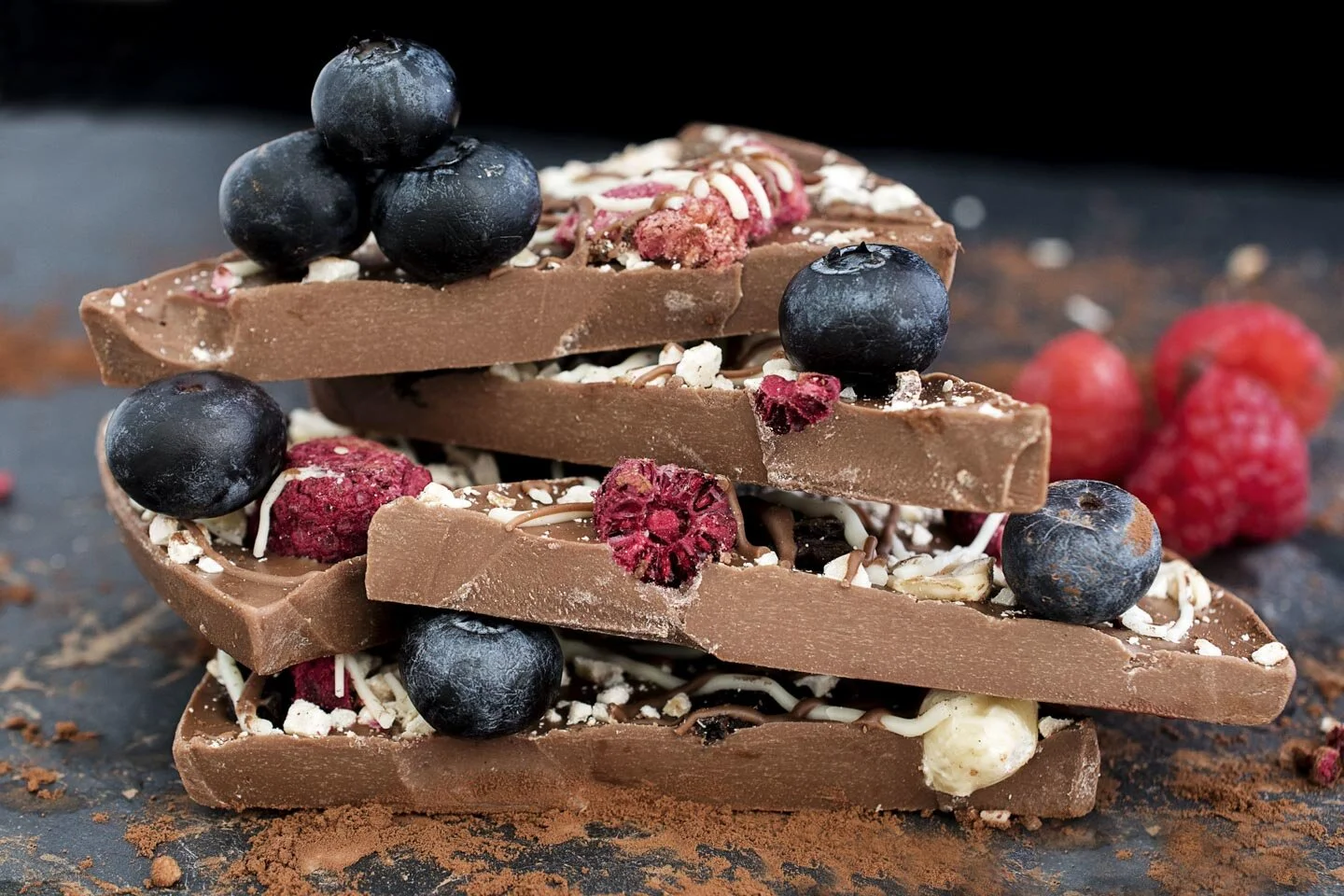 Stacked chocolate pieces with blueberries, raspberries, and nuts on a dark surface.