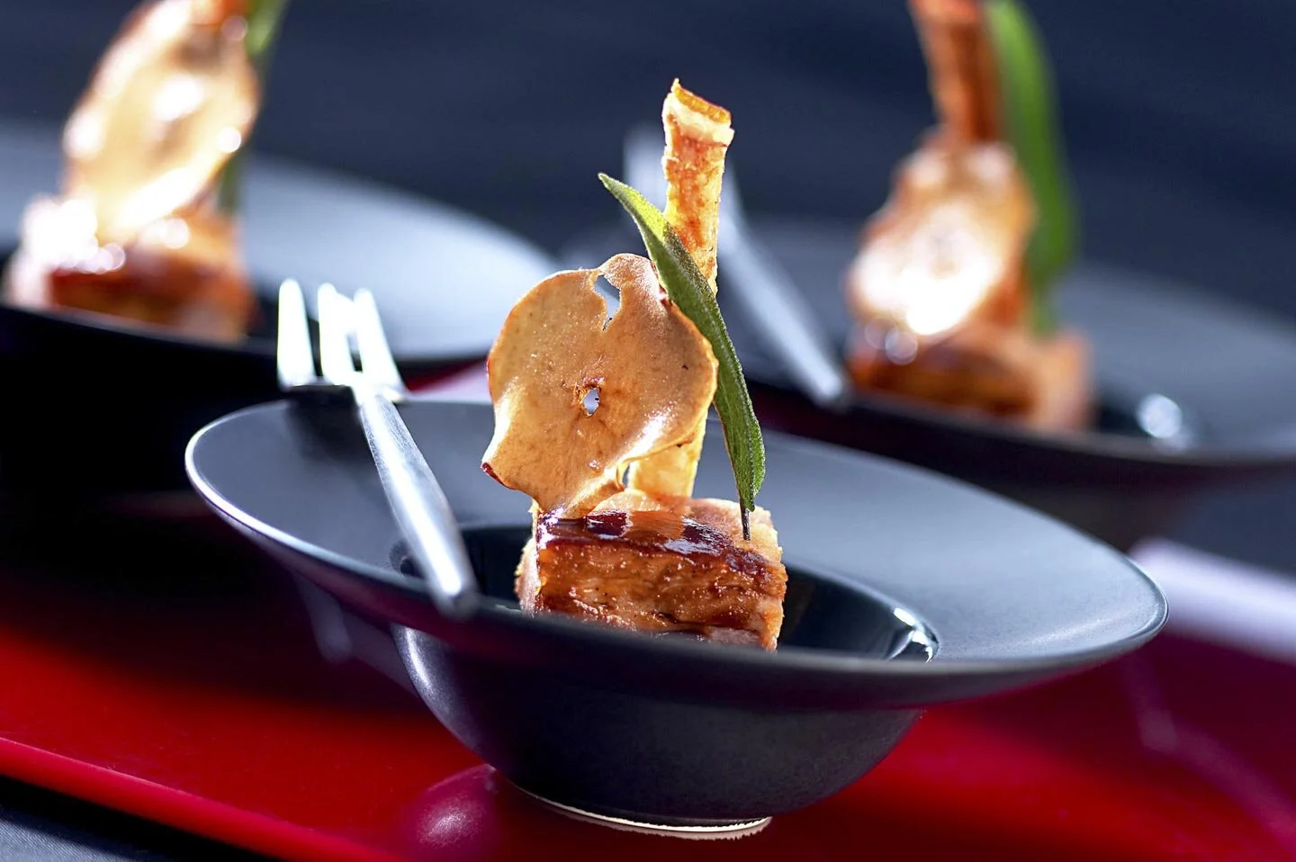 Gourmet appetizer in black bowl with caramelized meat, apple slice, and herb garnish