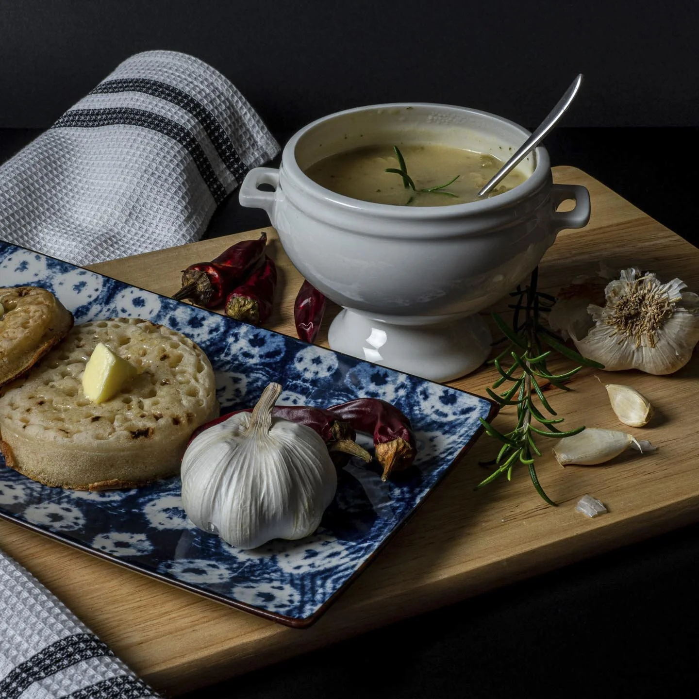 A bowl of soup with a sprig of rosemary on a wooden board, accompanied by a blue patterned plate holding crumpets with butter, garlic cloves, red chili peppers, and a white kitchen towel.