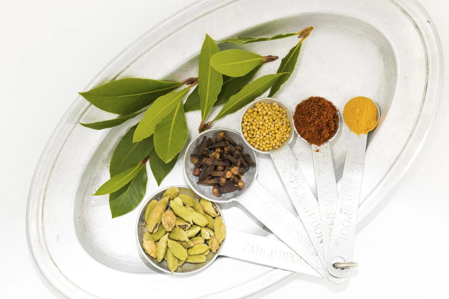 Assorted spices on a metal tray with bay leaves, cardamom pods, cloves, mustard seeds, ground chili, and turmeric in measuring spoons.