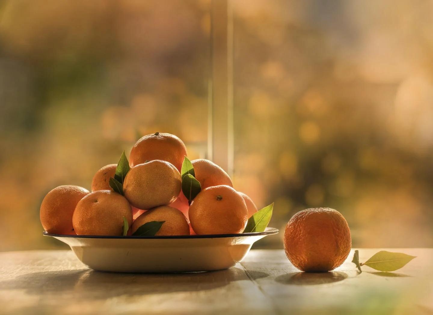 A bowl of ripe oranges with green leaves on a wooden table, lit softly by sunlight. A single orange with a leaf is placed adjacent to the bowl.