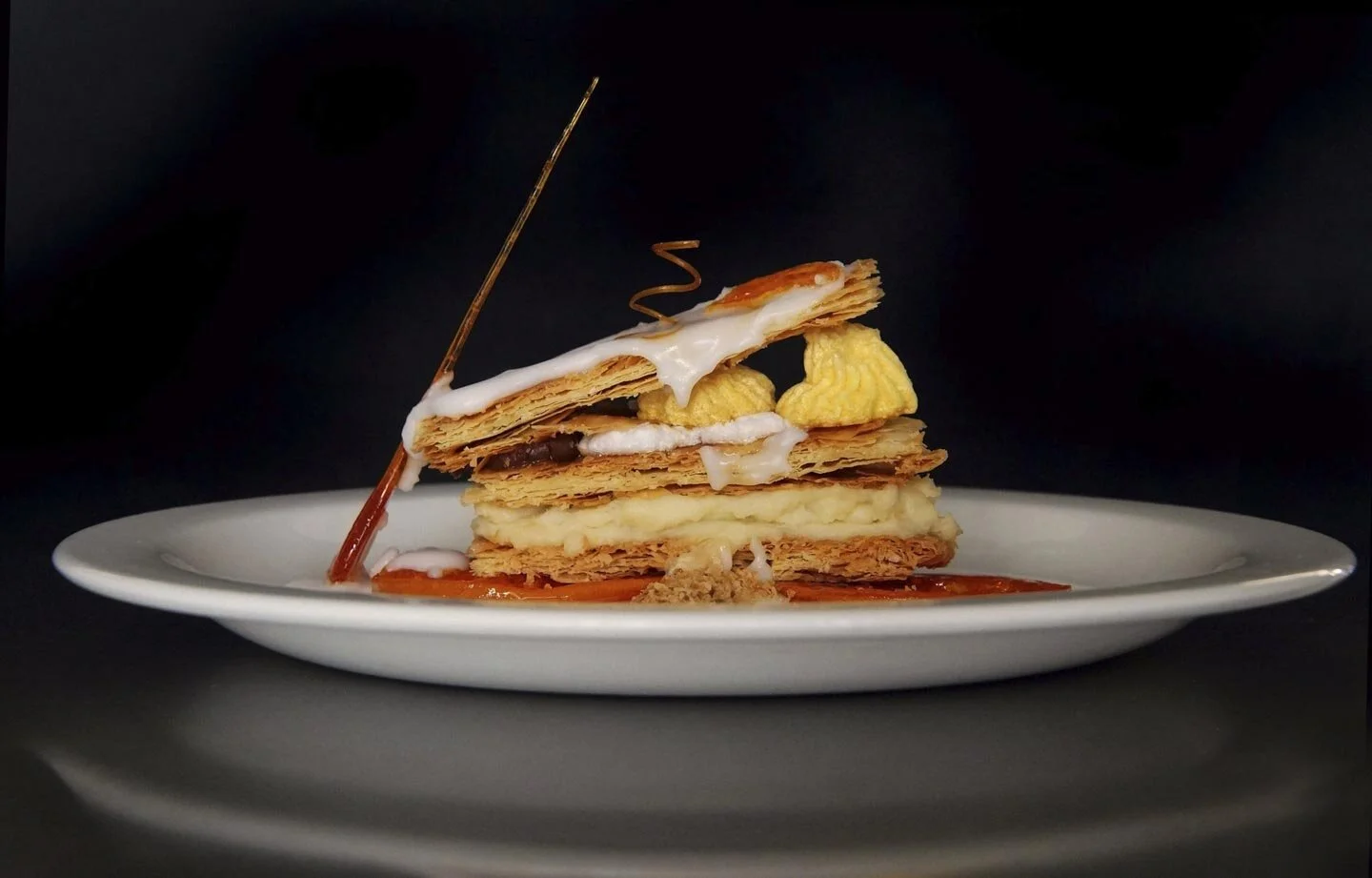 Elegant puff pastry dessert with cream, icing, and decorative caramel on white plate against dark background.