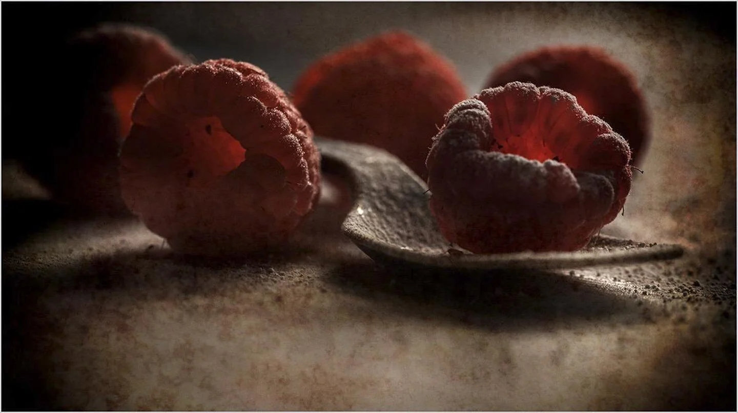 Close-up of ripe raspberries on a textured surface with dramatic lighting, shallow depth of field highlighting one raspberry on a spoon.