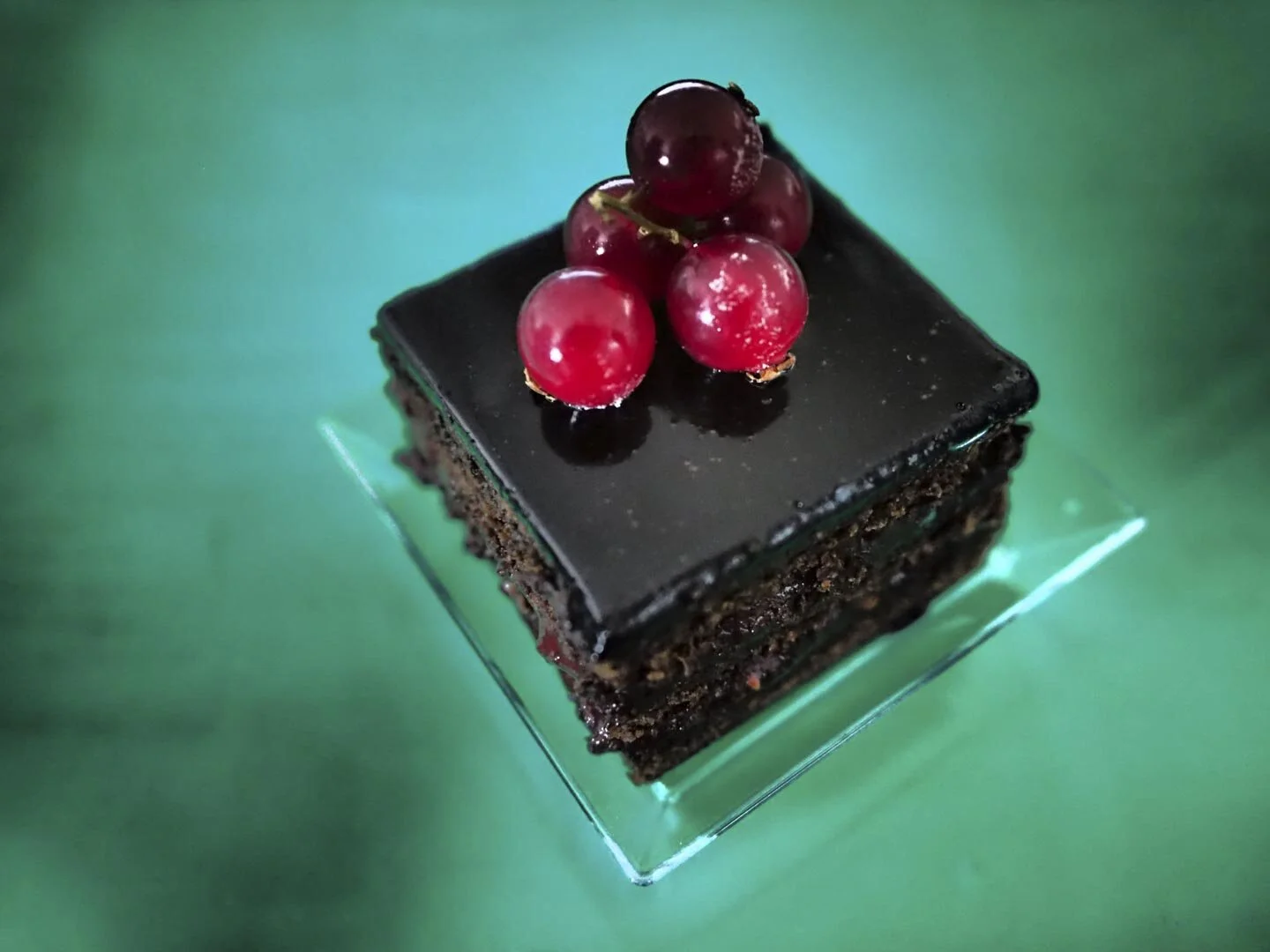 Chocolate cake slice with glossy frosting and red currants on top, placed on a glass dish.
