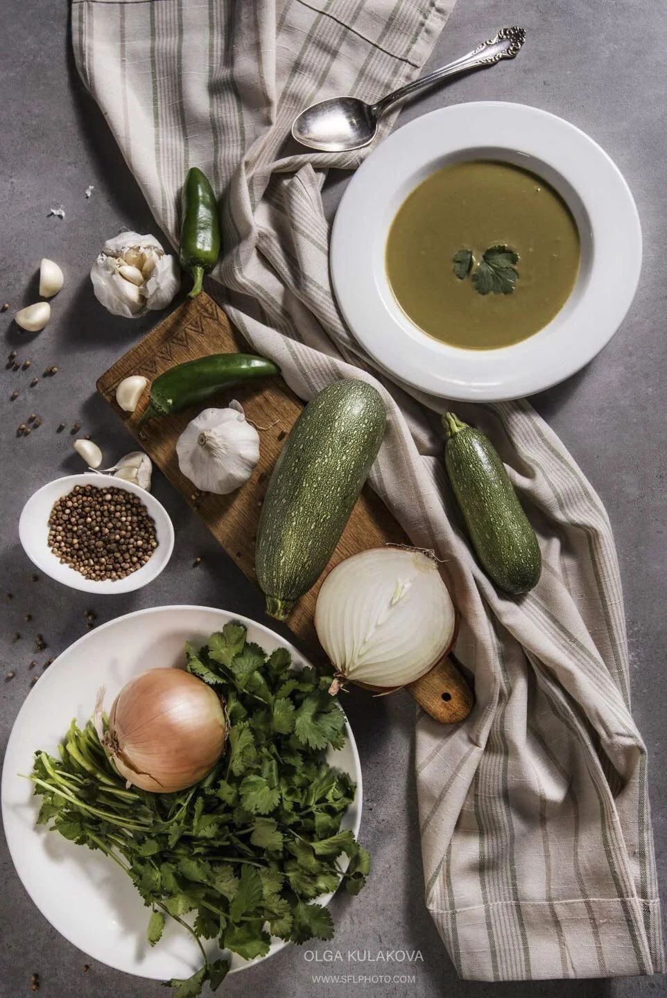 Plate of green soup with garnish, surrounded by ingredients including garlic, zucchini, jalapeños, onions, cilantro, and coriander seeds on a gray surface with a striped cloth.