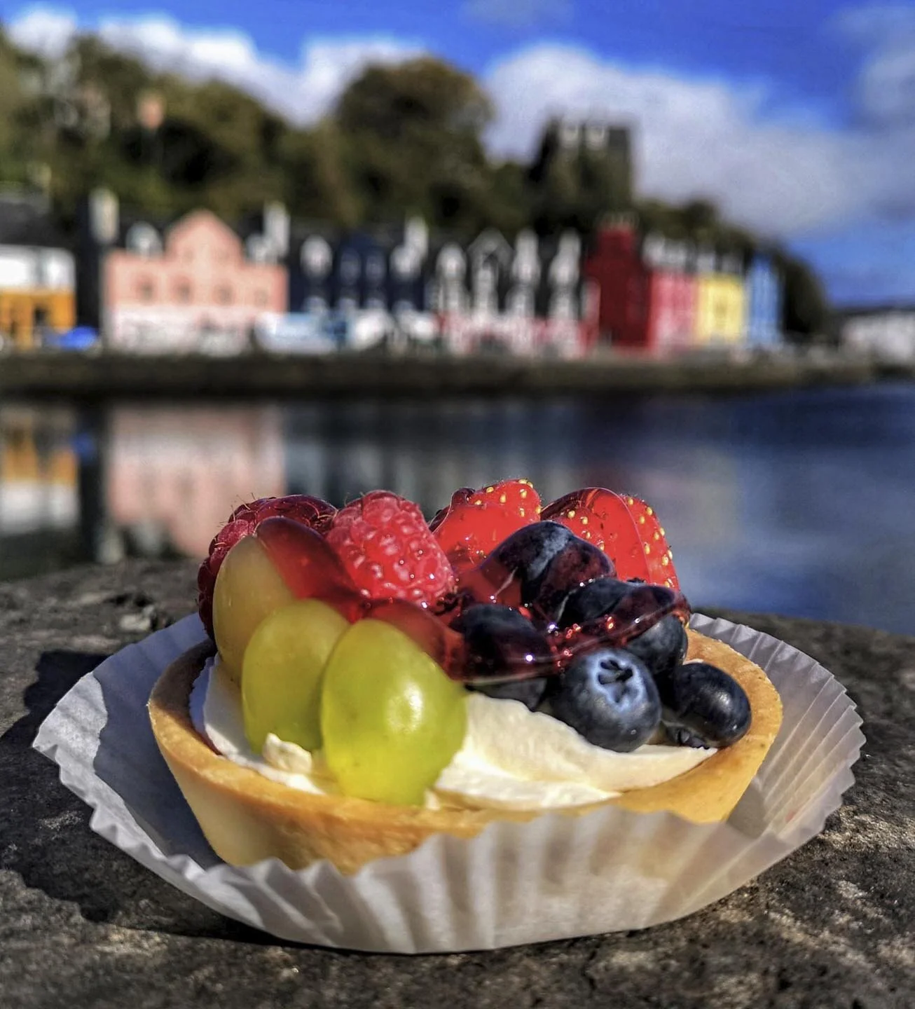Fruit tart with grapes, blueberries, raspberries, and strawberries on pastry shell, with blurred colorful buildings in background.