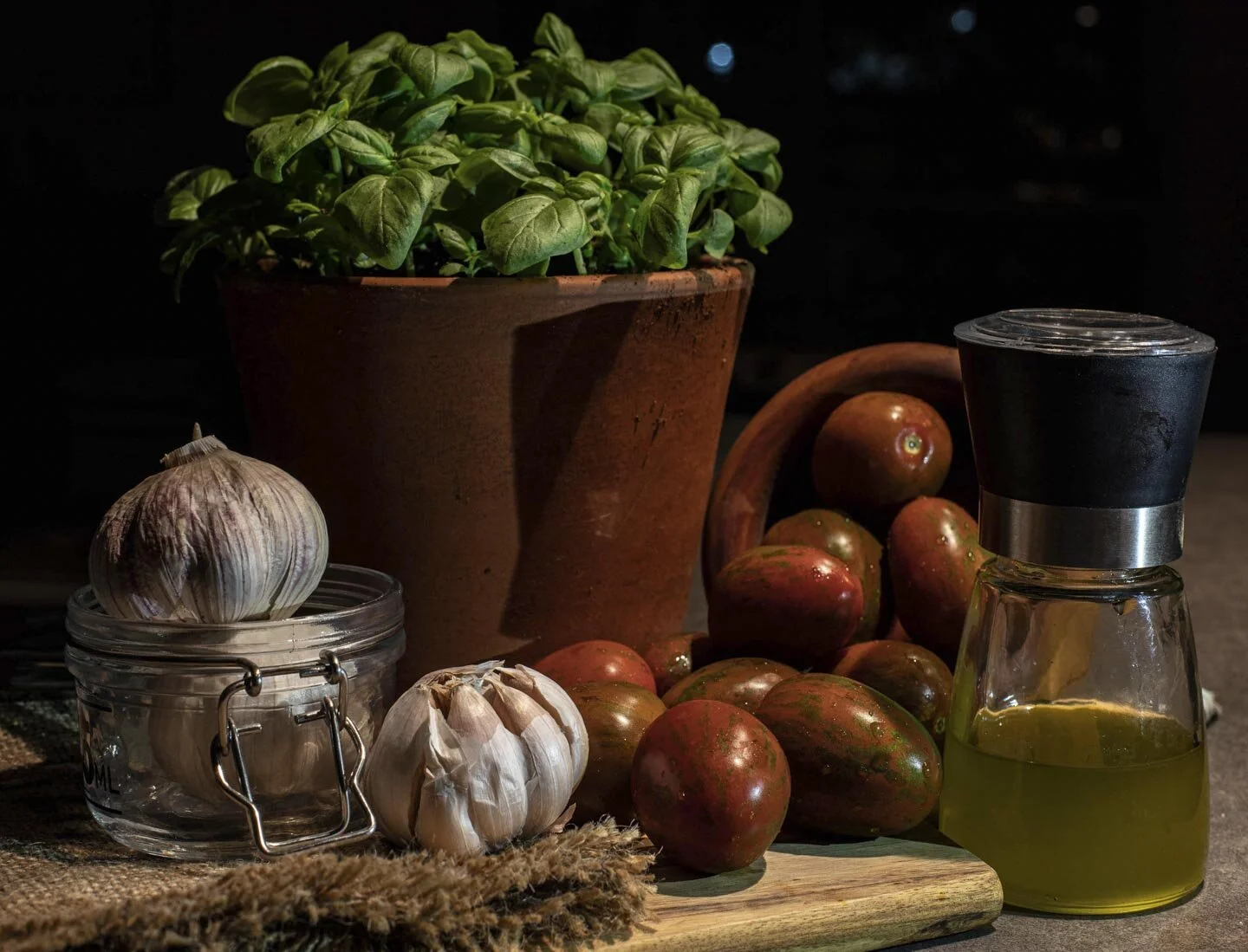 A rustic kitchen setting featuring a pot of fresh basil, a garlic bulb on a glass jar, another garlic bulb on the countertop, ripe tomatoes spilling from a wooden bowl, and a glass bottle of olive oil with a black cap.
