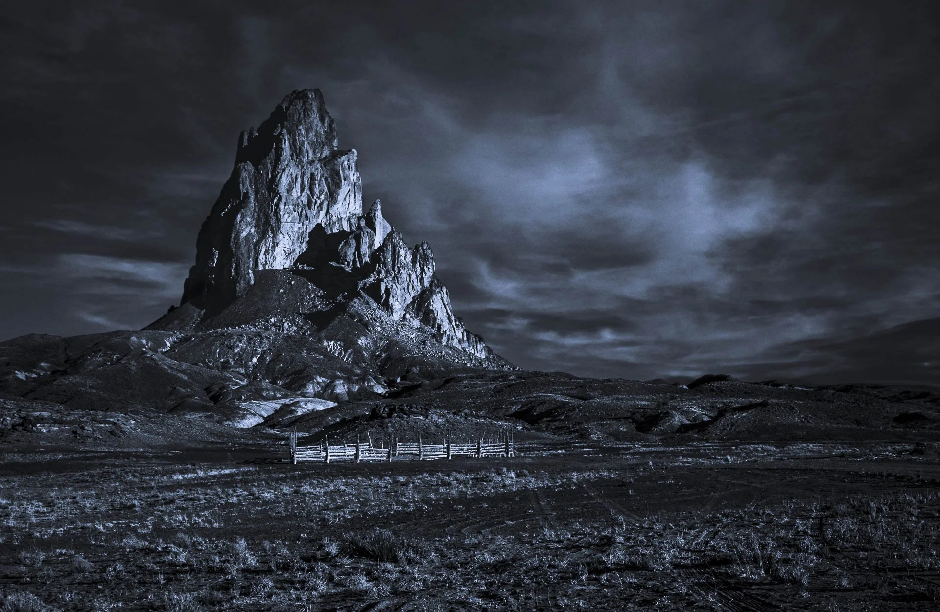 Monochrome image of a rugged mountain peak under cloudy skies, with dry, barren land in the foreground.