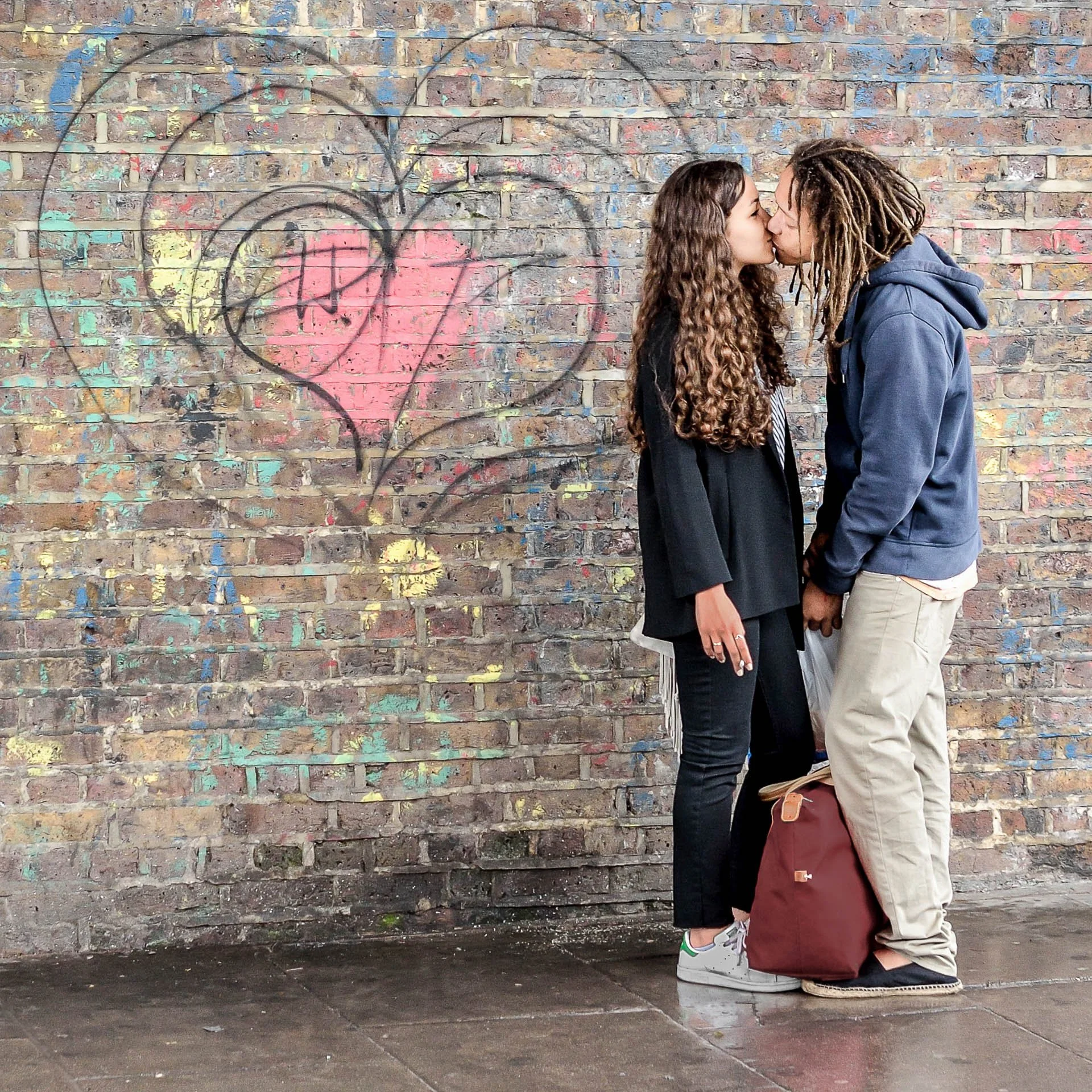 A couple kissing in front of a brick wall with heart graffiti.