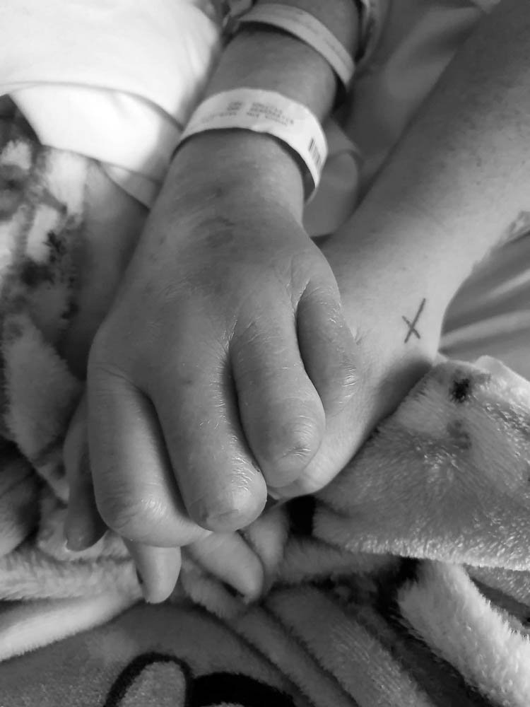 Two hands holding, one with a hospital wristband, on a patterned blanket.