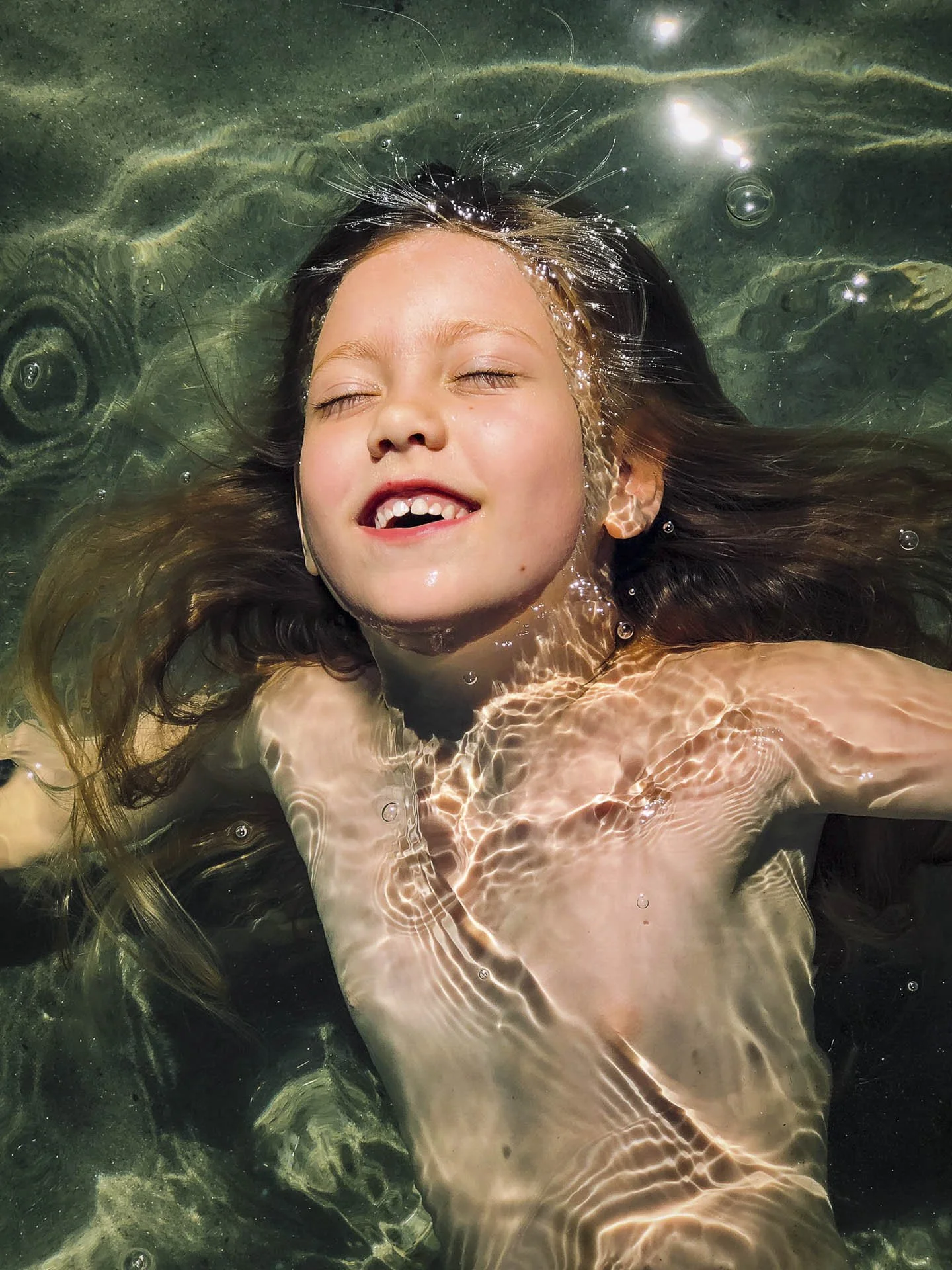 Child floating and smiling with closed eyes in clear water, sunlight creating patterns on the skin.