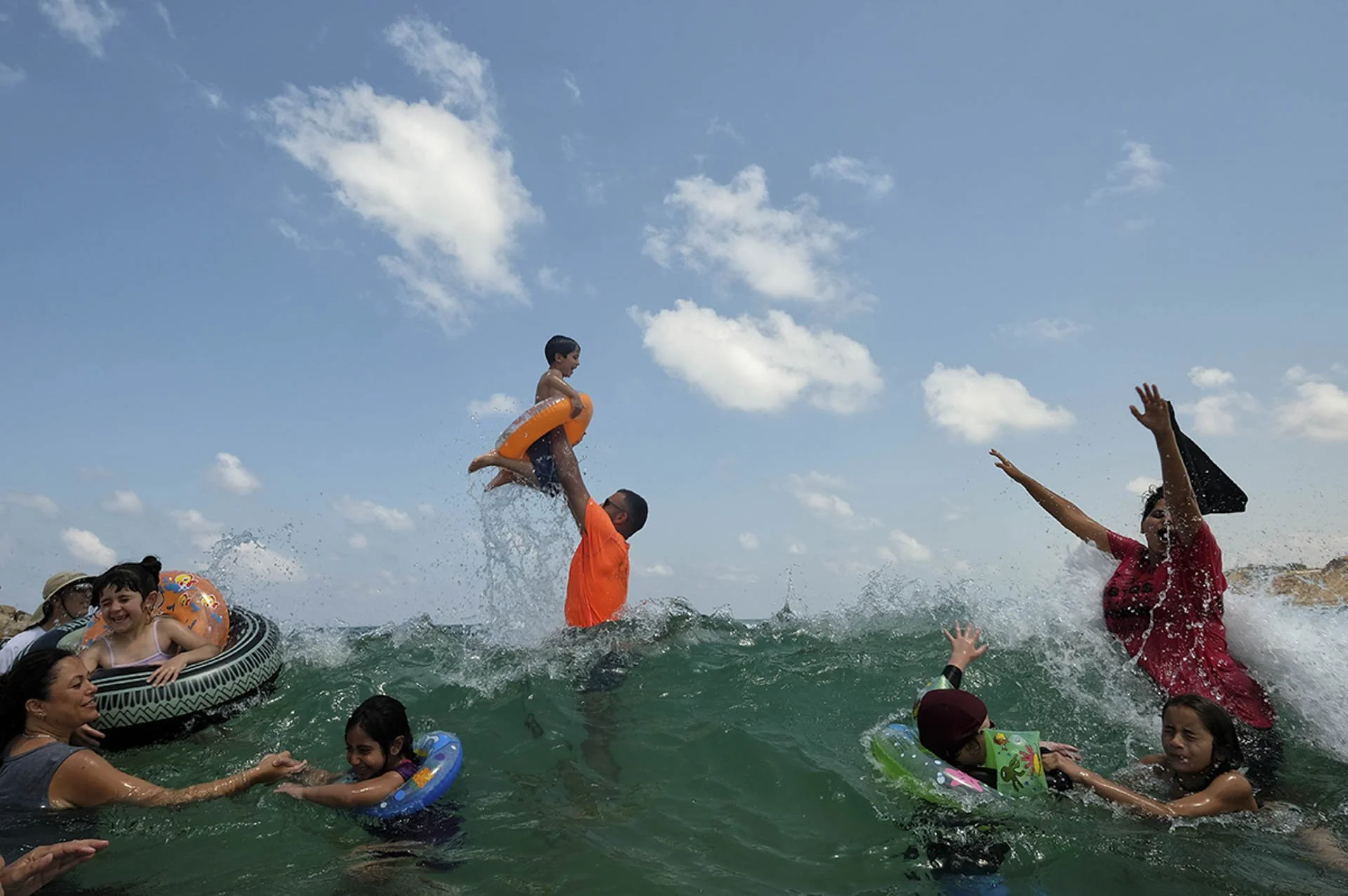 People enjoying a day at the beach, with children in swim rings and an adult lifting a child in the water.