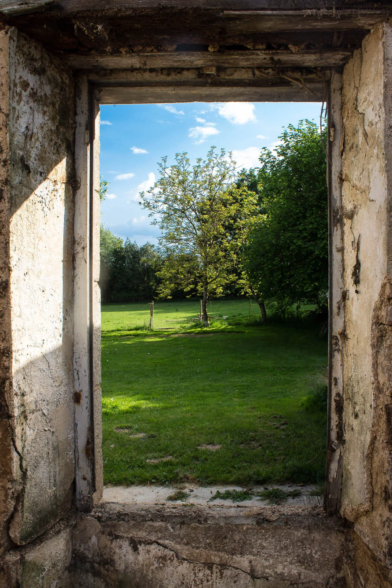View through an old stone window frame revealing a grassy lawn and trees under a blue sky.