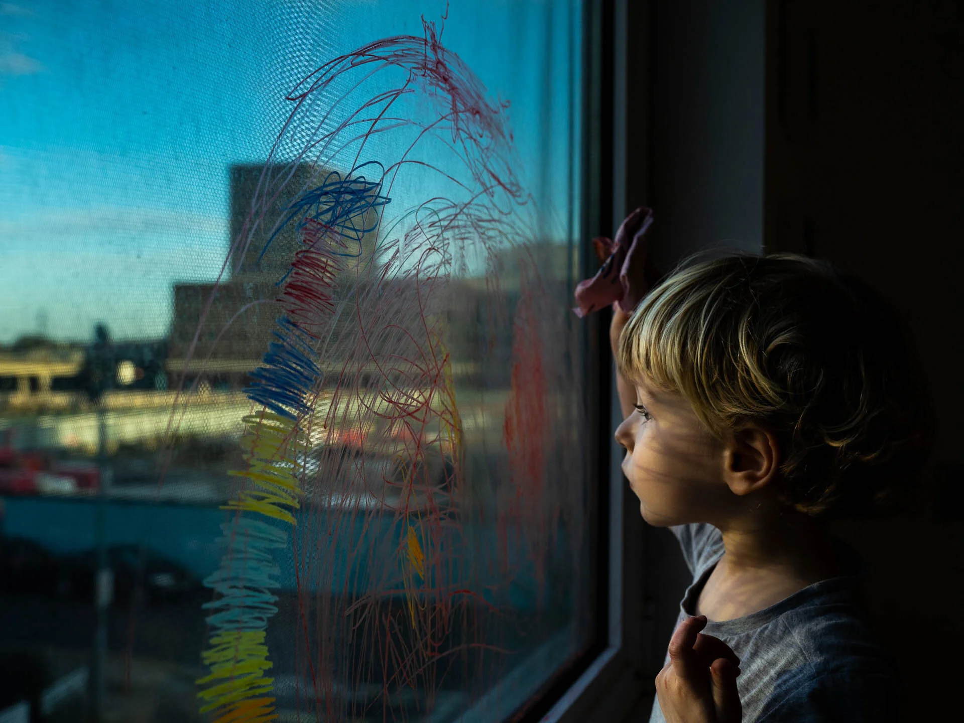 Child looking out window with colorful scribbles on glass, cityscape in background.