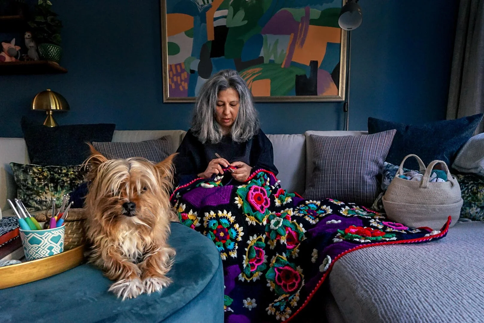 Woman with gray hair crocheting a colorful blanket on a sofa, with a small dog sitting nearby on an ottoman in a cozy living room.