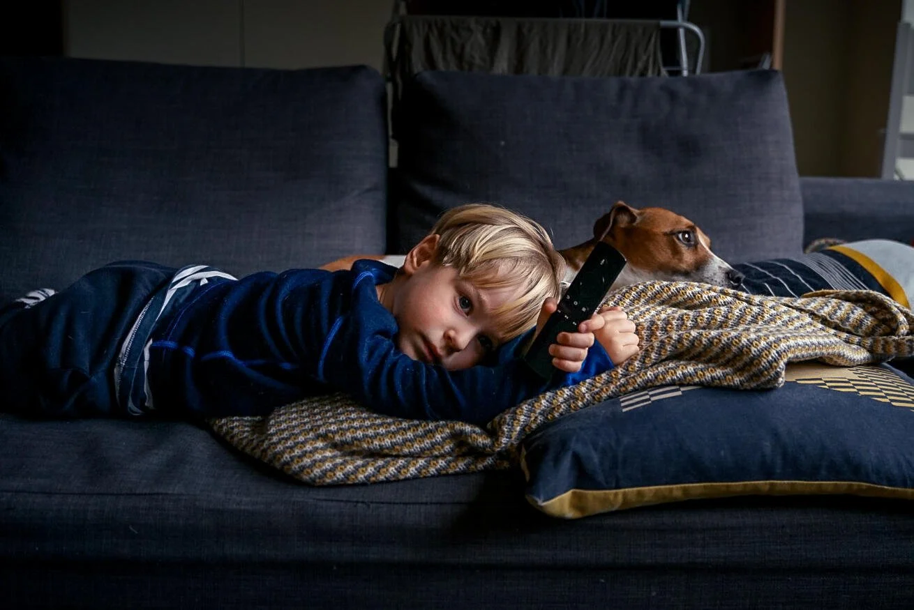 A young child holding a remote control lies on a couch next to a brown and white dog, both resting on a patterned blanket.