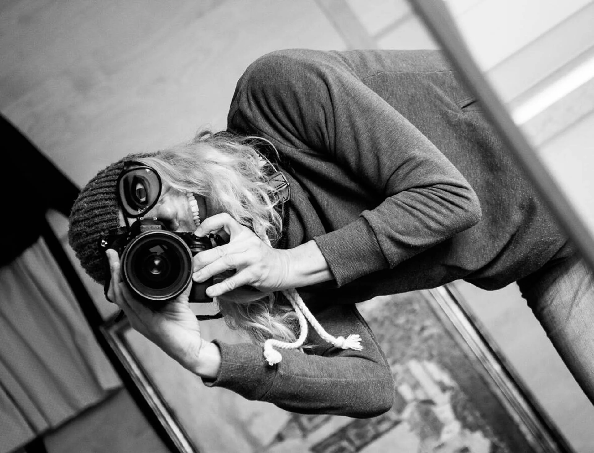 Person wearing a hat and goggles taking a photo with a camera in black and white.