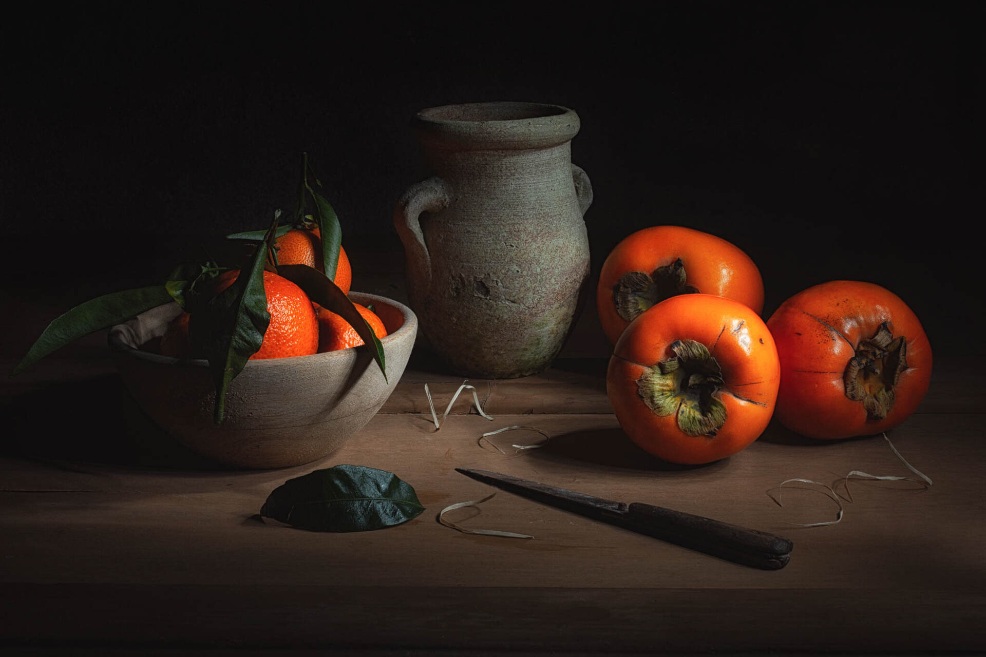Still life with a bowl of tangerines, persimmons, an earthenware jug, a knife, and a leaf on a wooden table.