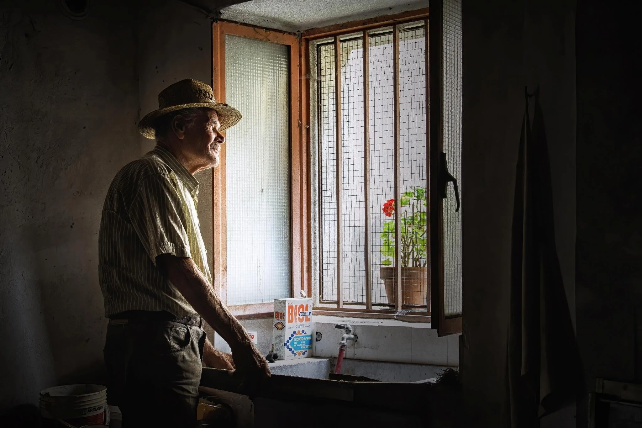 Elderly man in straw hat looking through window with potted red flowers on ledge, standing in dimly lit room.