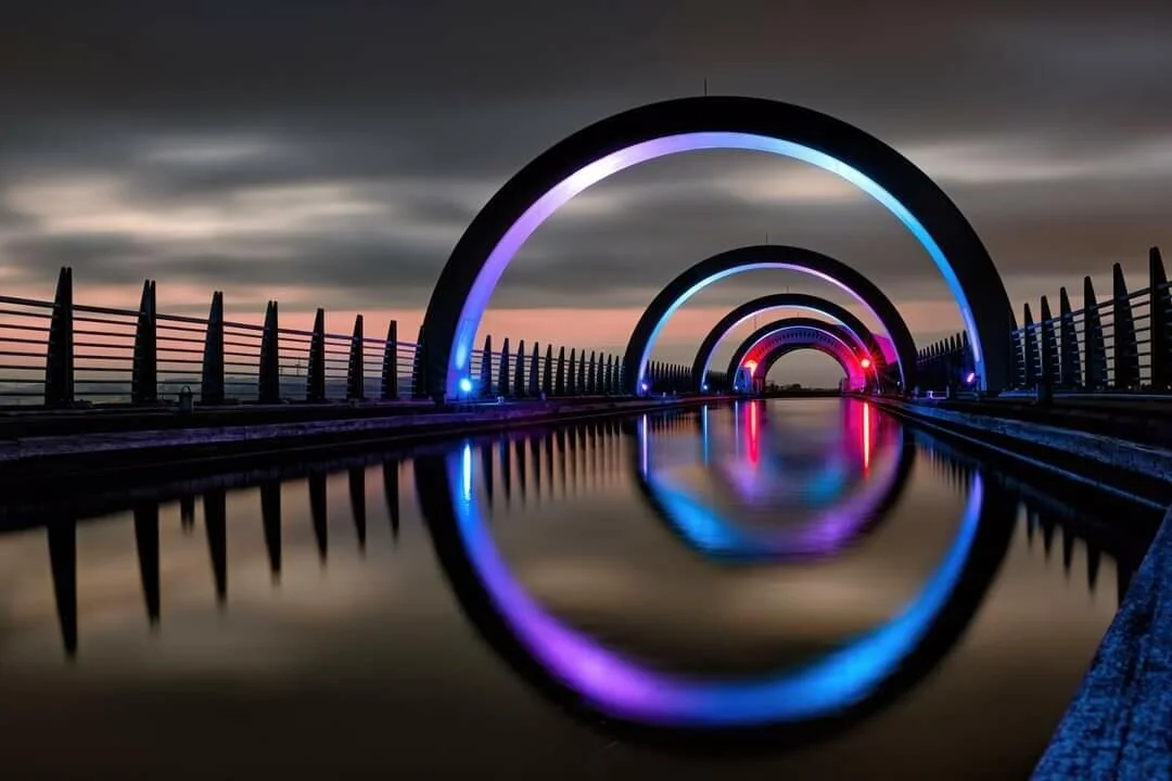 Illuminated aqueduct with colorful arches at night, reflecting on still water.
