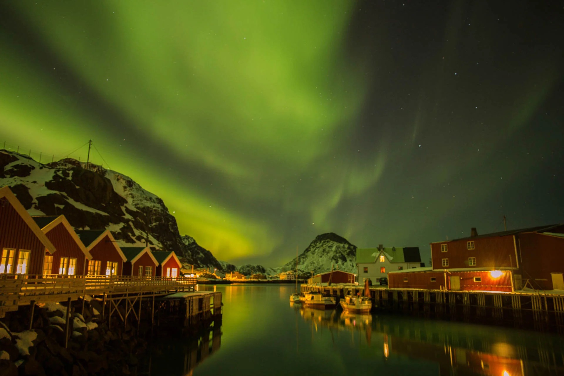 Scenic view of the Northern Lights over snow-covered mountains and wooden houses reflected in a calm body of water at night.