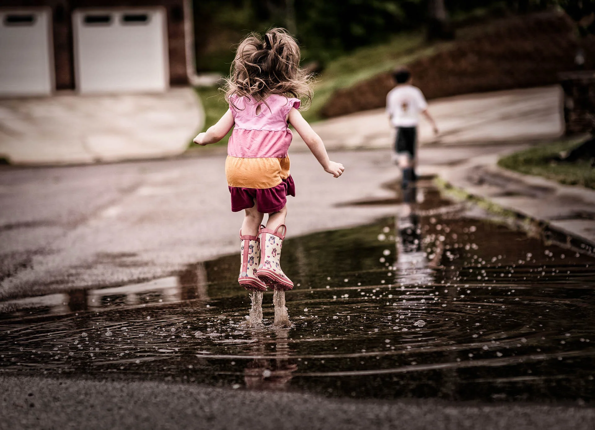 Child in pink rain boots jumping in a puddle on a suburban street.