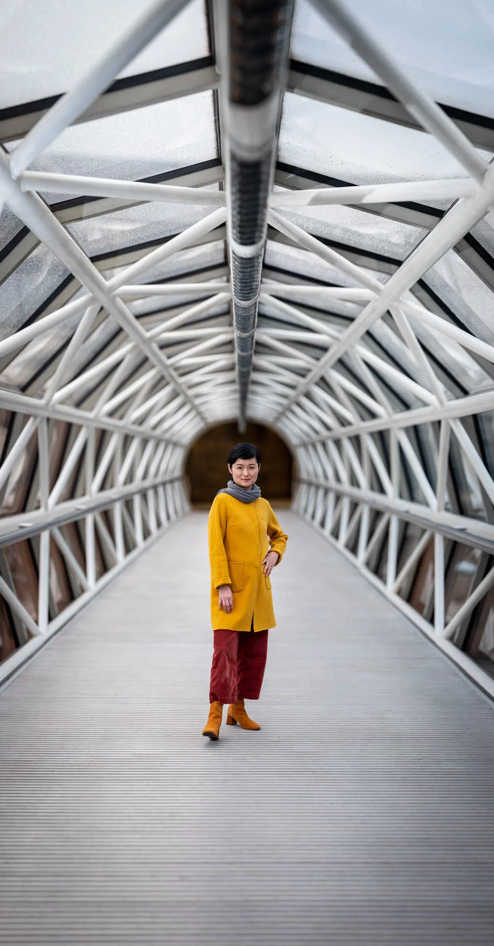 Person in yellow coat and red pants standing in a modern curved metallic tunnel.
